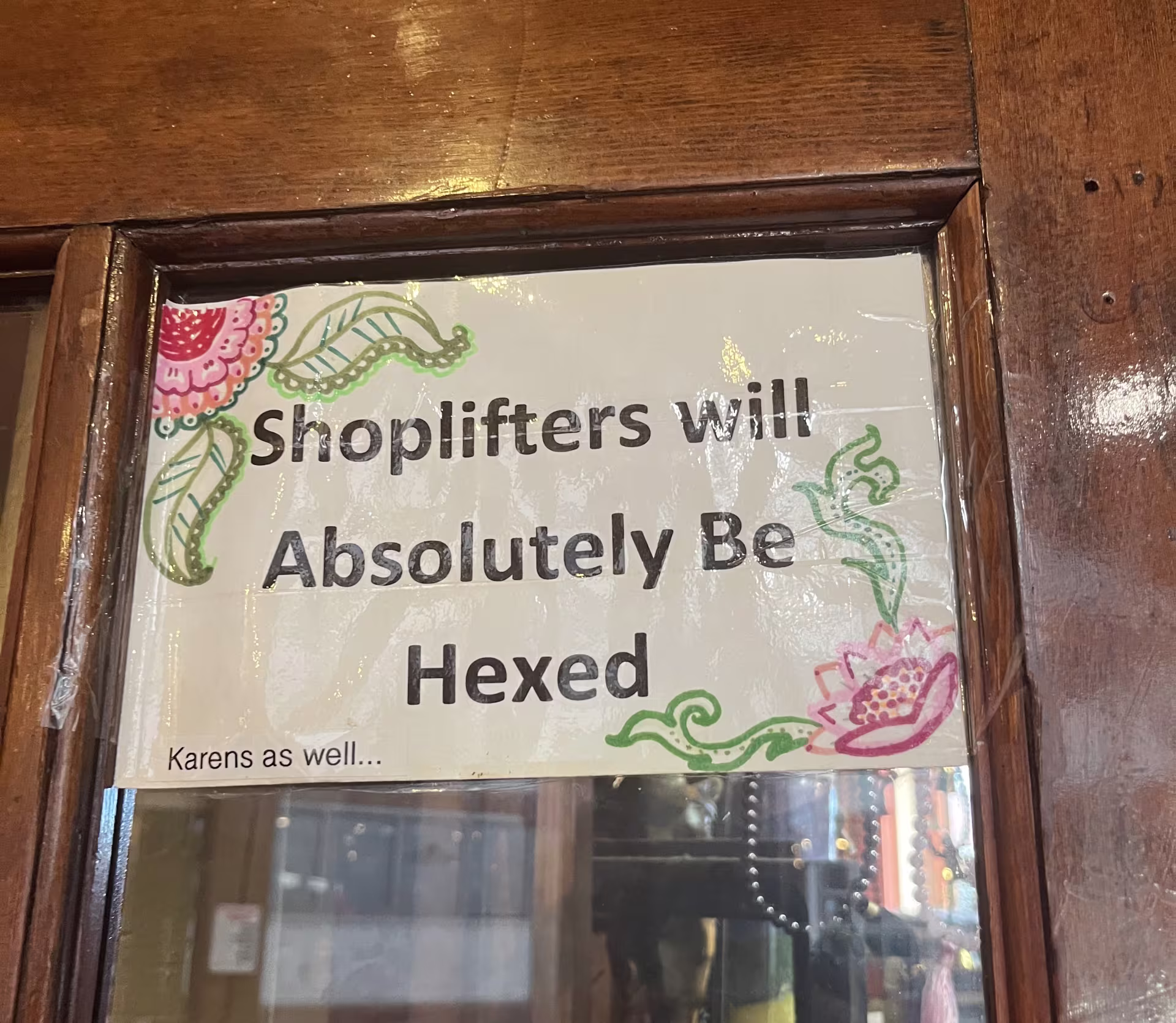 A sign with the text "Shoplifters will Absolutely Be Hexed" surrounded by drawings of flowers. In the corner is smaller text reading "Karens as well..."