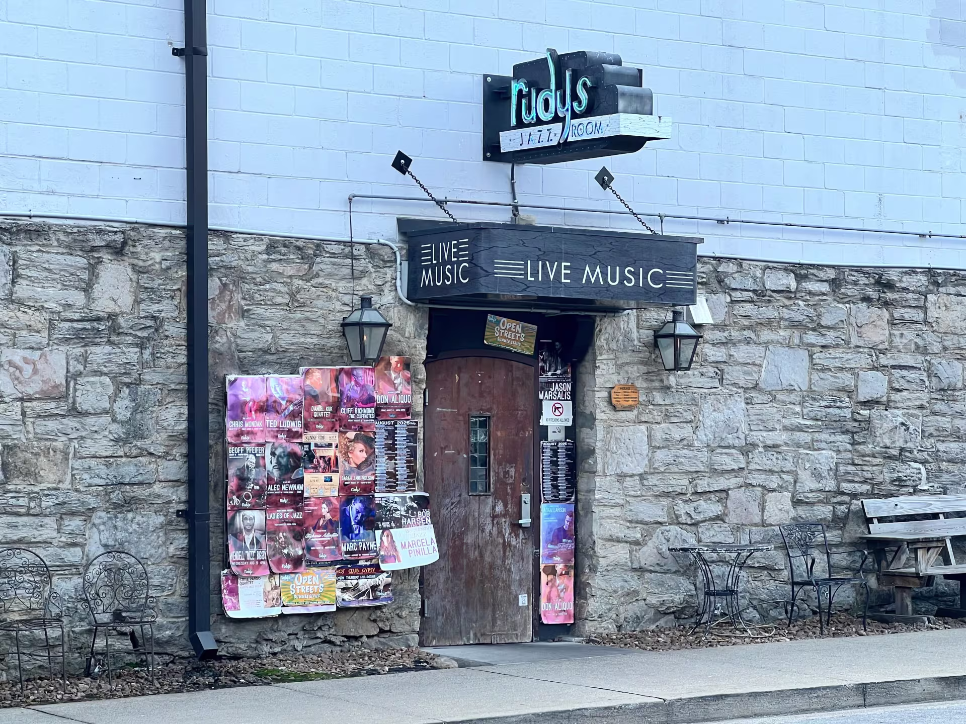 A stone and white brick building with a bulletin board packed with paper fliers to the left of a weathered wooden door. Above the door is a black awning that says "Live Music," and a black sign that says "rudy's jazz room" in teal lettering.