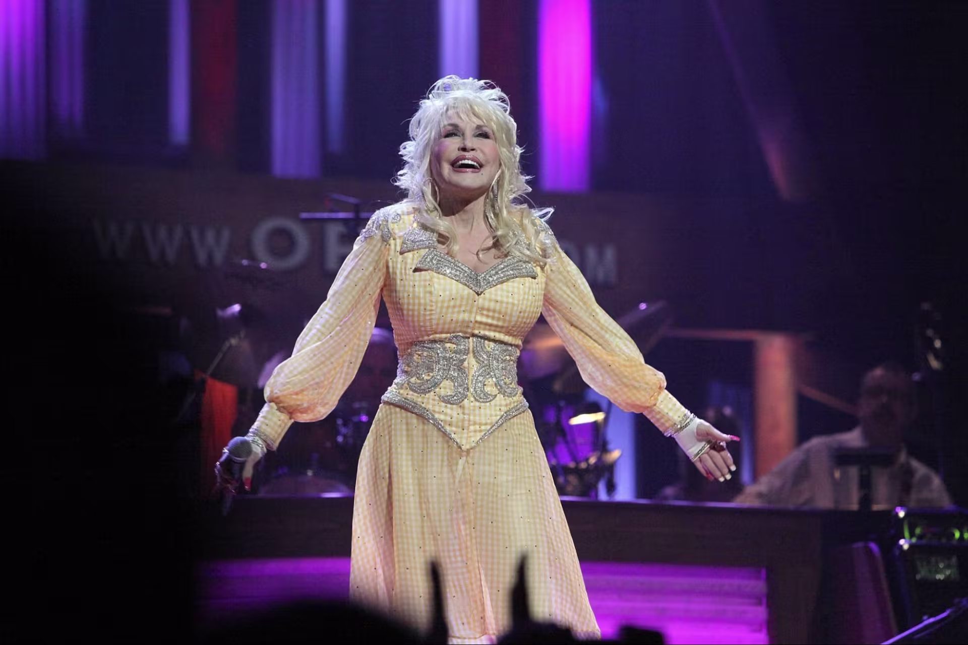  Dolly Parton stands on the Opry stage in front of its iconic backdrop, which is lit in purple. She is standing with her arms outstretched and a microphone in her right hand. She is wearing a yellow gingham dress with silver rhinestone details, and smiling broadly while looking up at the balcony.