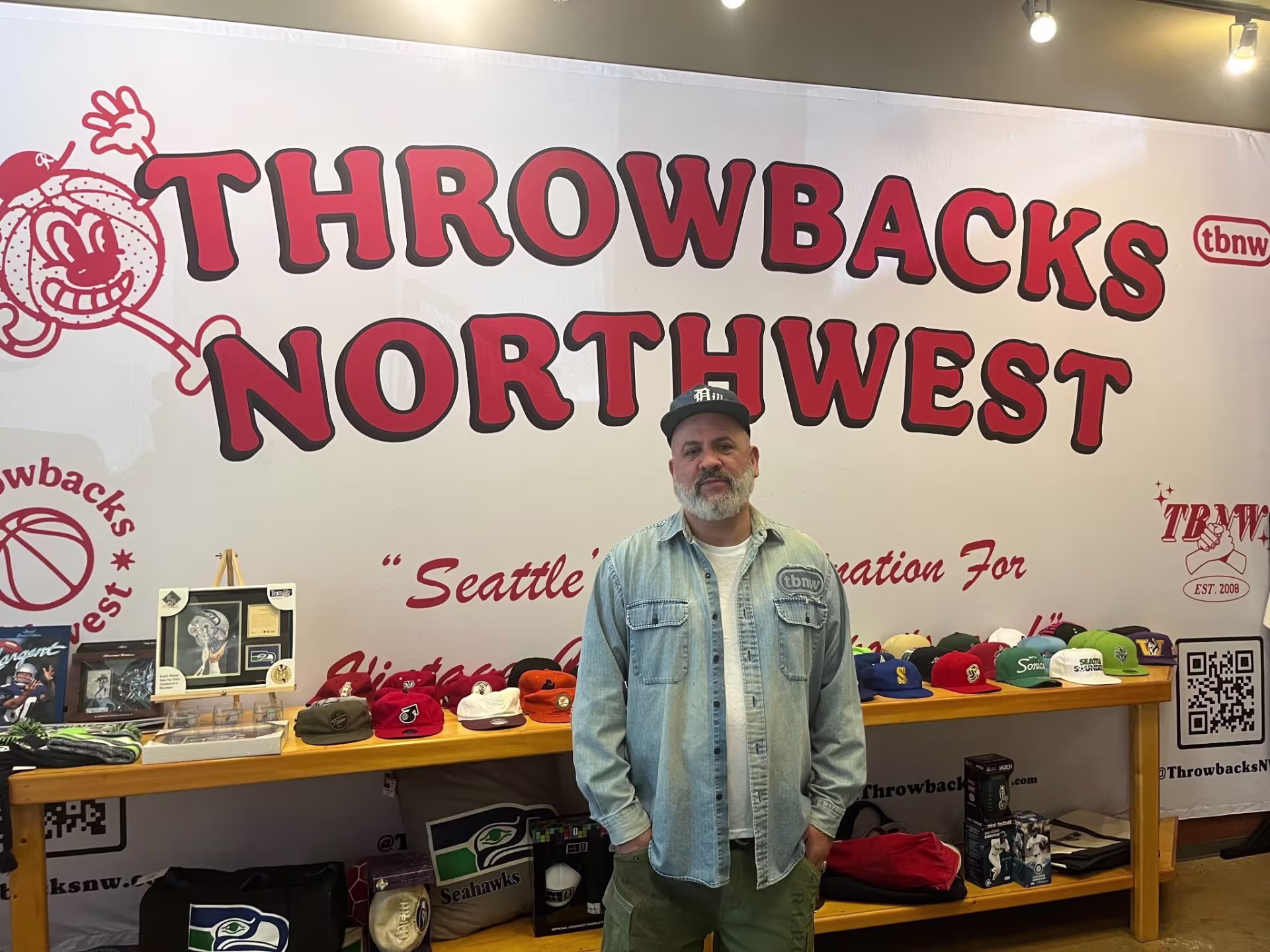 A man standing in front of an assortment of sports hats, seahawks pillows and other gear. He is also standing in front of a sign that says "throwbacks northwest" 