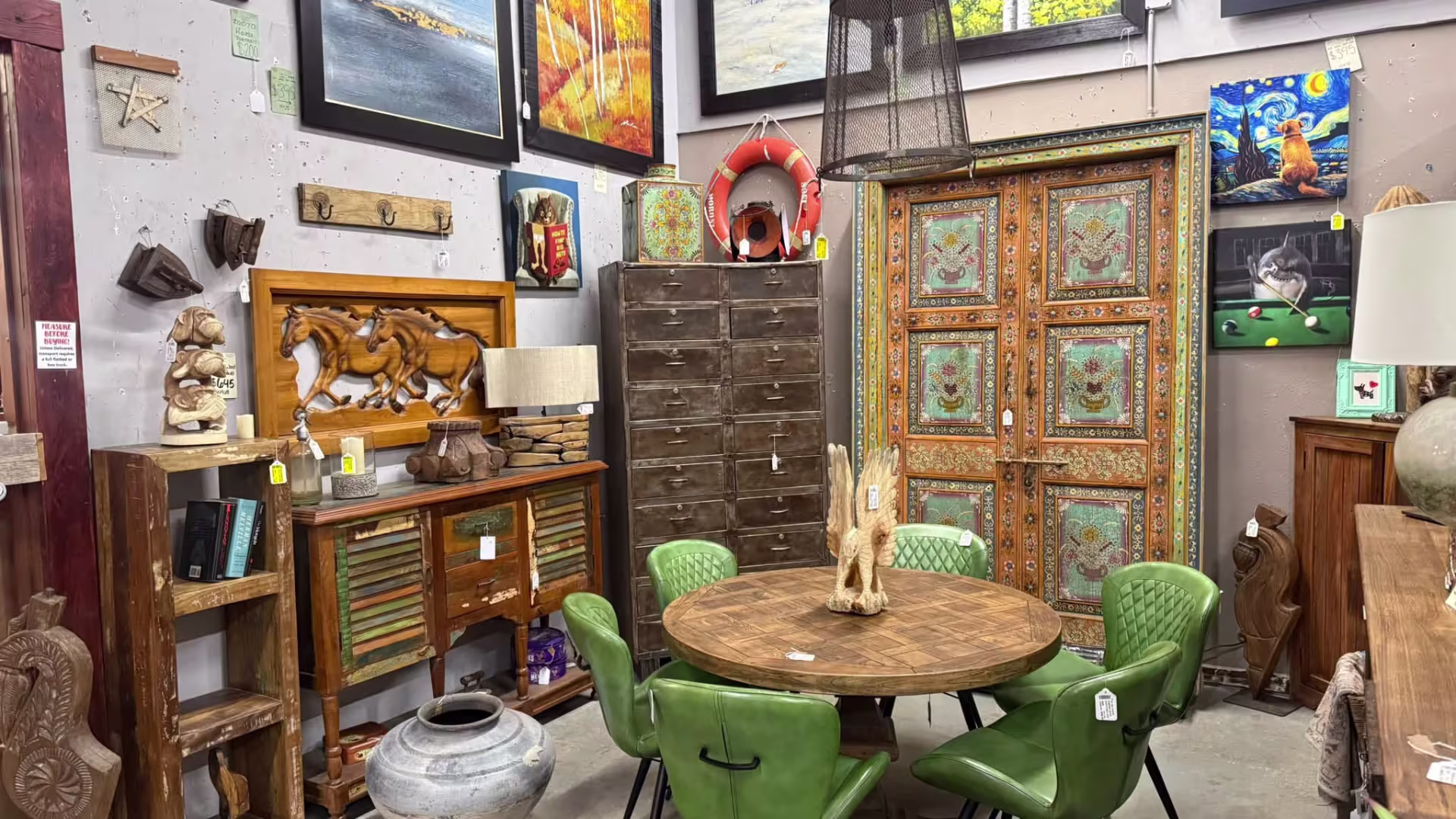 Green chairs around a wooden table with an ornate wardrobe in the background and art on the walls.