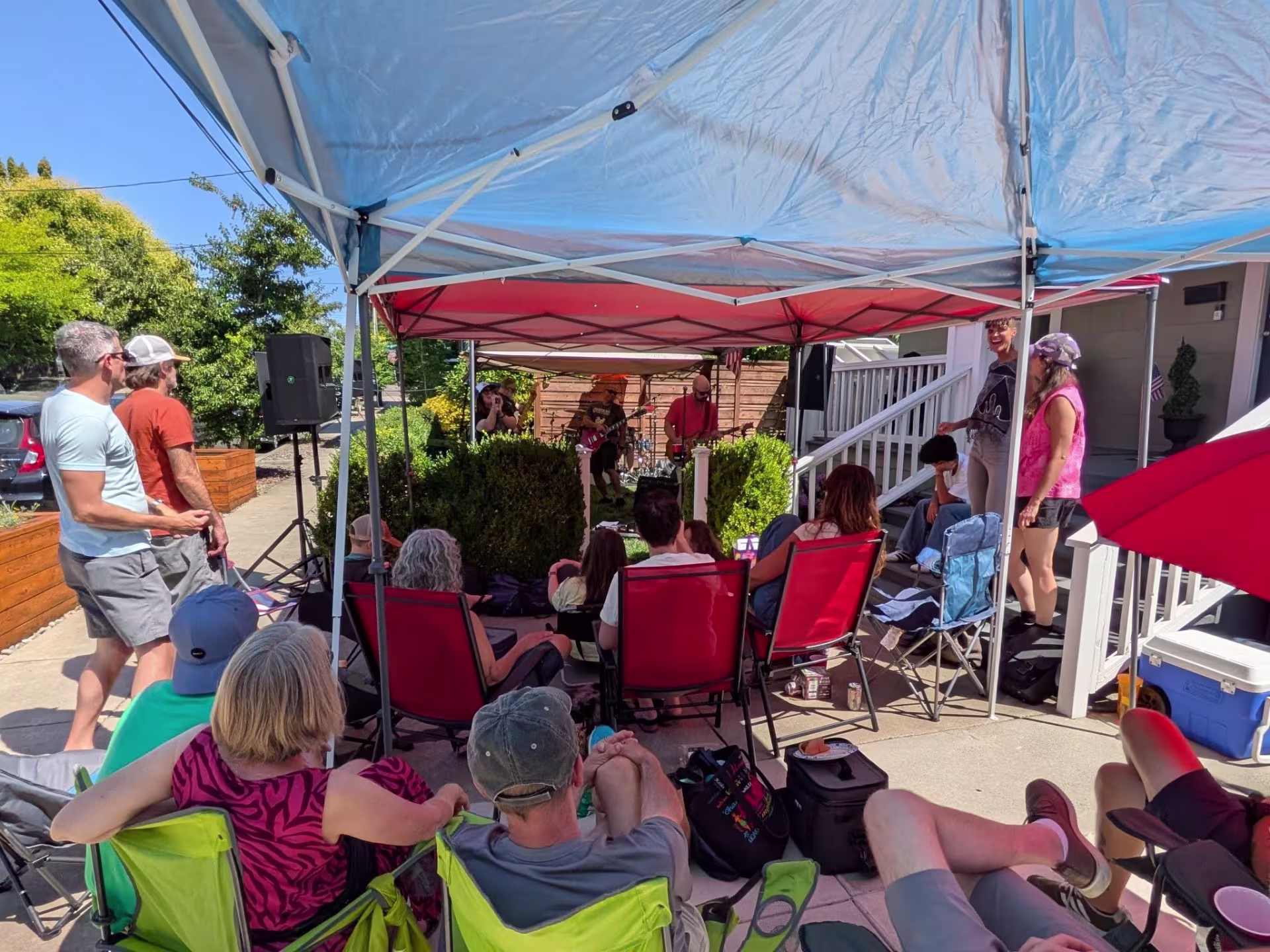 People sit in camp chairs underneath pop-up canopies in a driveway while listening to musicians play guitar in the front yard.