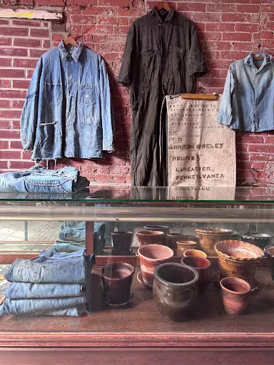 A display at Meadowsweet General Store in Norris Square featuring vintage denim workwear and clothing alongside handmade ceramic pots, arranged in front of a weathered brick wall.