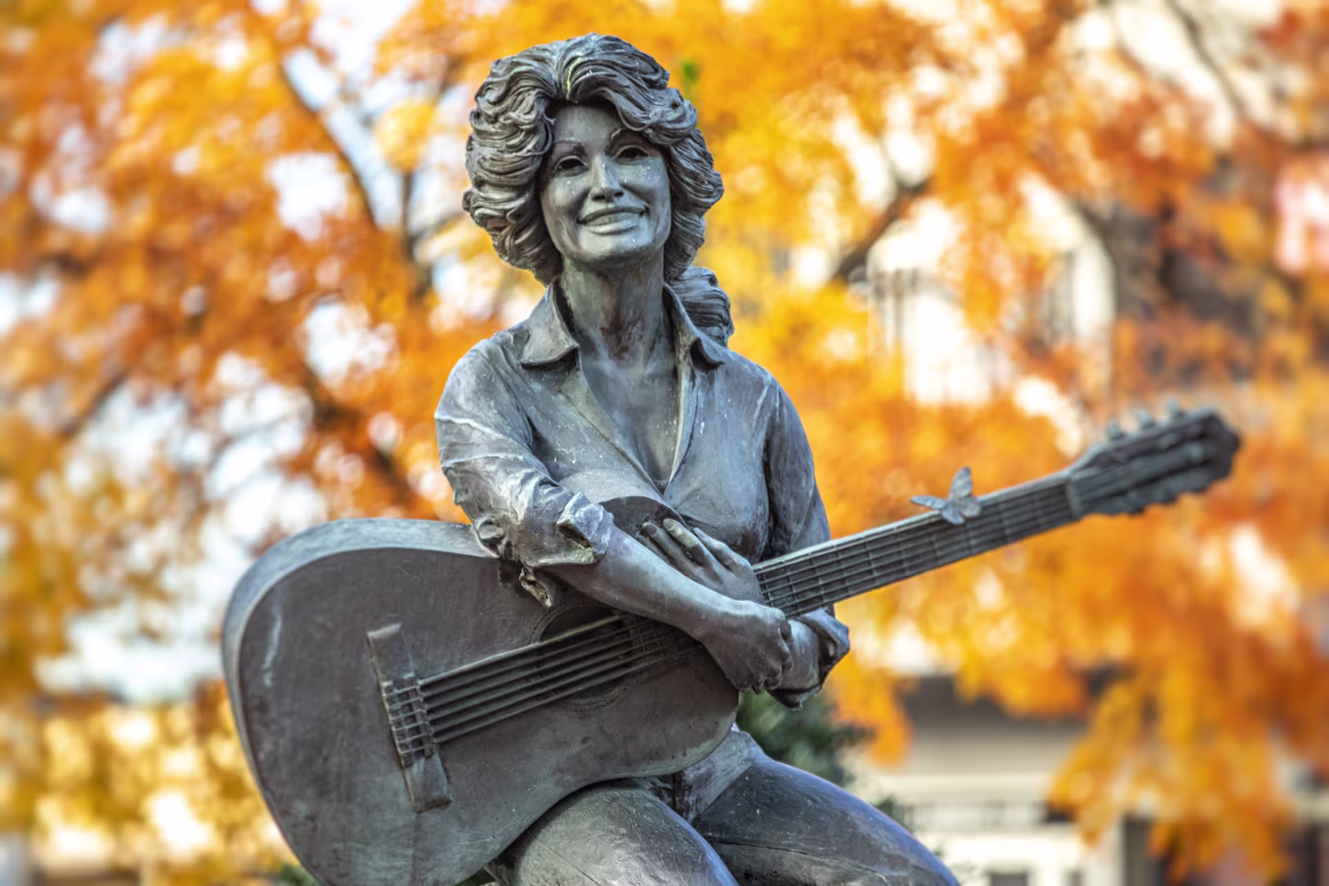 A statue of Dolly Parton holding a guitar with a butterfly on it, with trees that have orange leaves in the background.