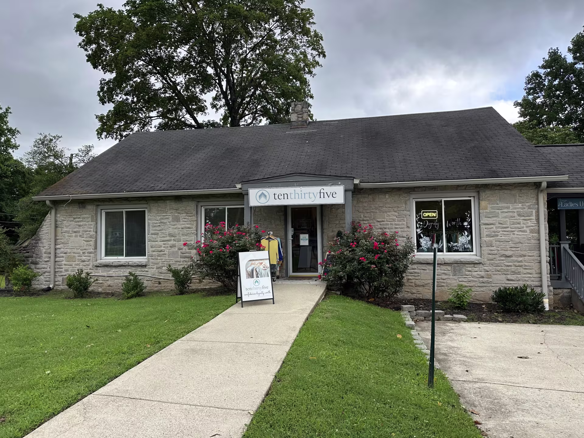 A stone cottage viewed from the front. There is green grass alongside a concrete sidewalk and driveway leading to the cottage. A sign above the door reads "tenthirtyfive." Behind the house are trees and a blue sky clouded with gray.