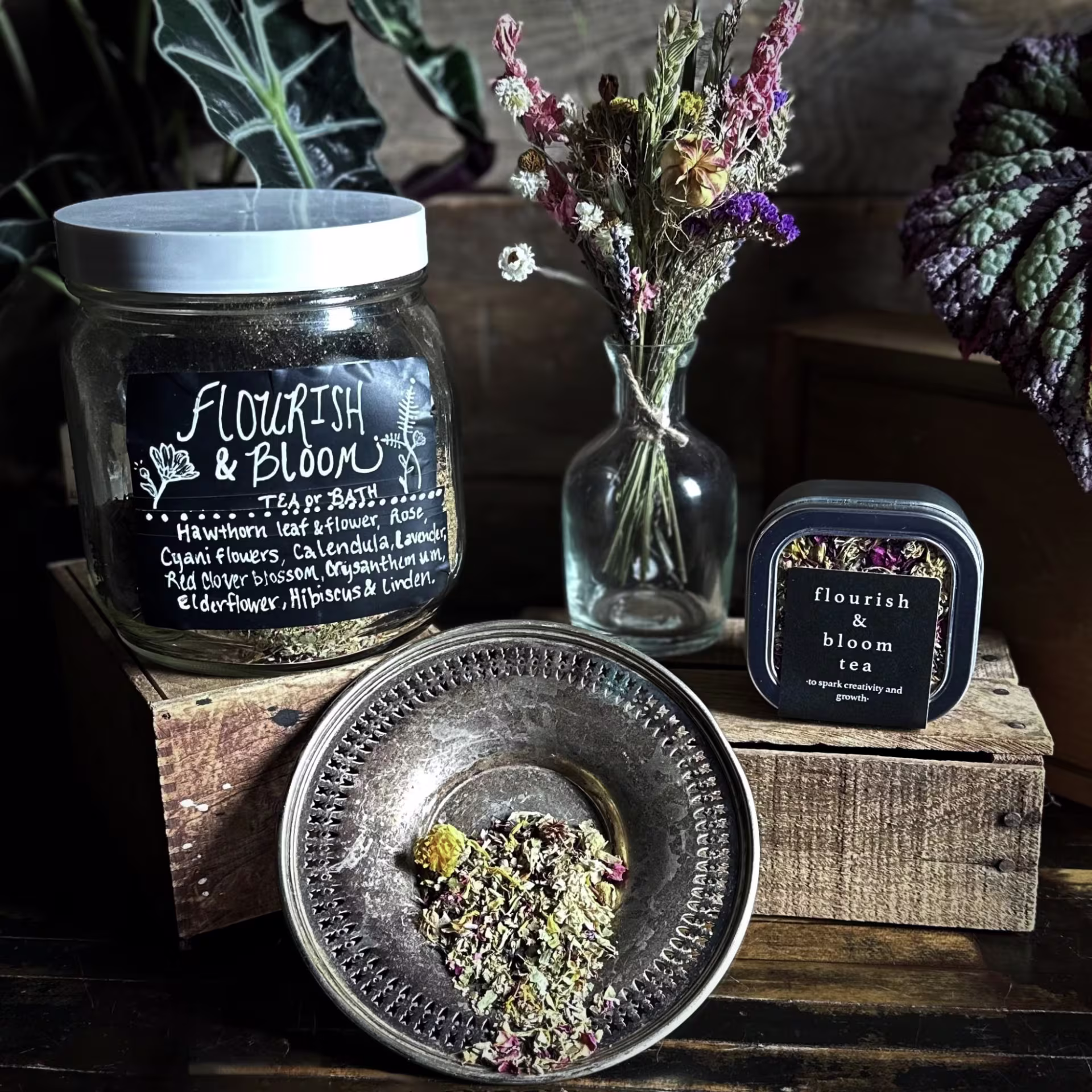Handmade tea in a jar with a white lid, in a silver bowl, and in a small package arranged on a wooden table and risers with a dried bouquet of flowers in the background.