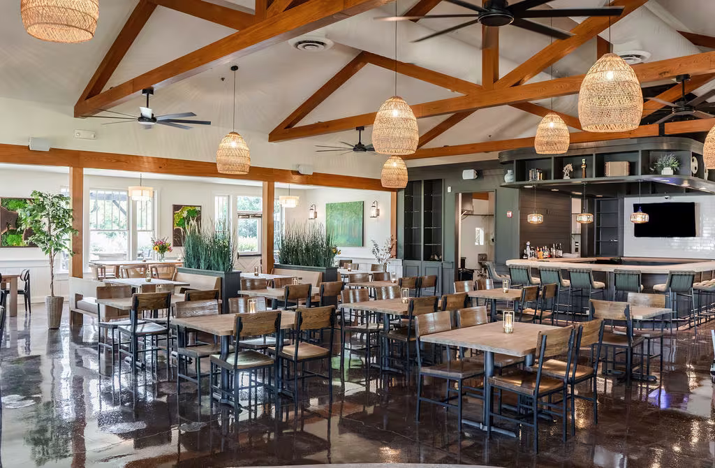 A restaurant with wooden beams on a tall ceiling, white walls, and wooden tables and chairs. Woven light fixtures hang from the ceiling.