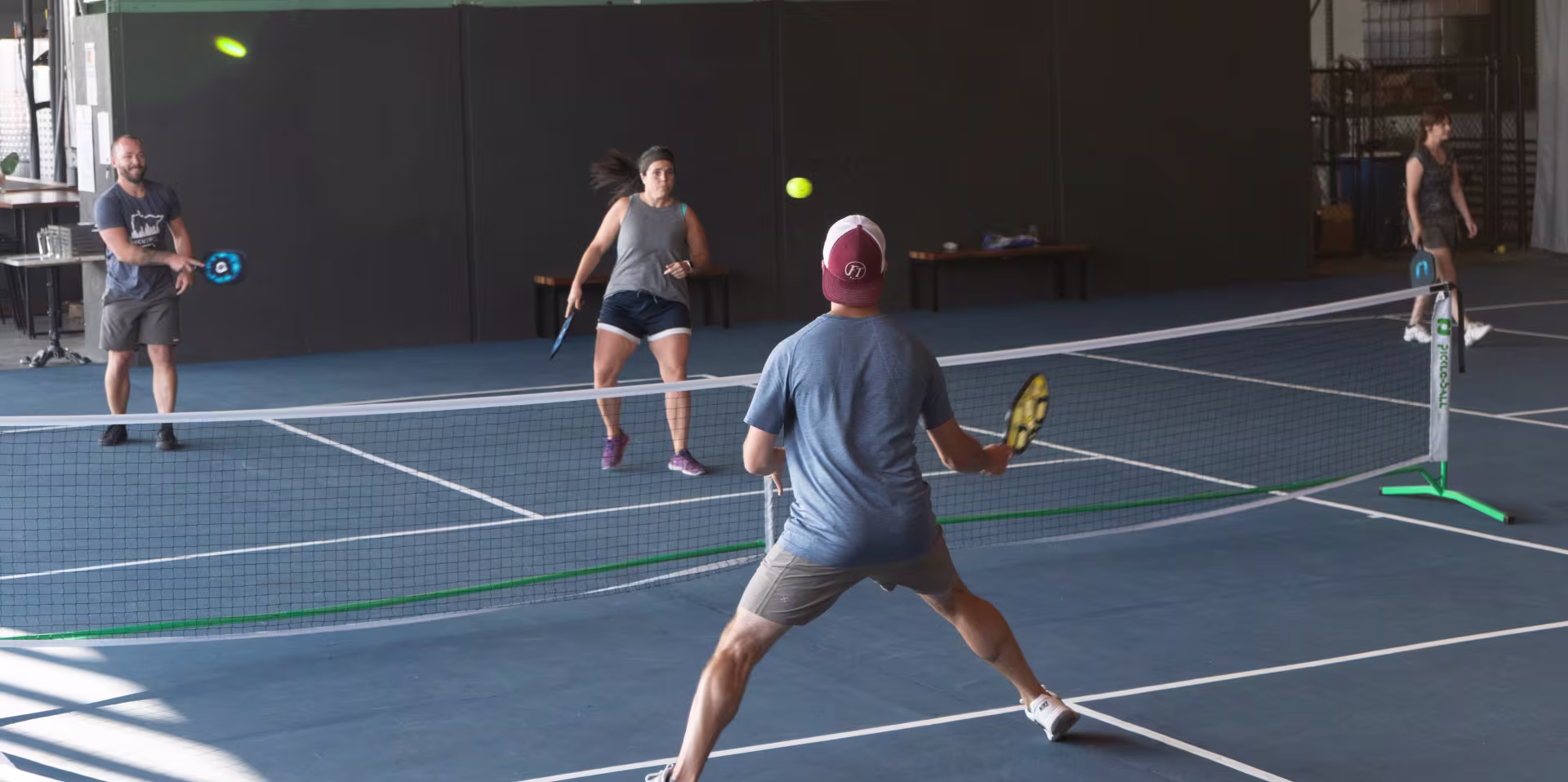 Three players are engaged in an active pickleball game and stand in different parts of the indoor court. One has his back to the camera and holds a paddle in his right hand oriented towards a ball that is mid-air. A woman and a man play across from him while another player walks on another court nearby.