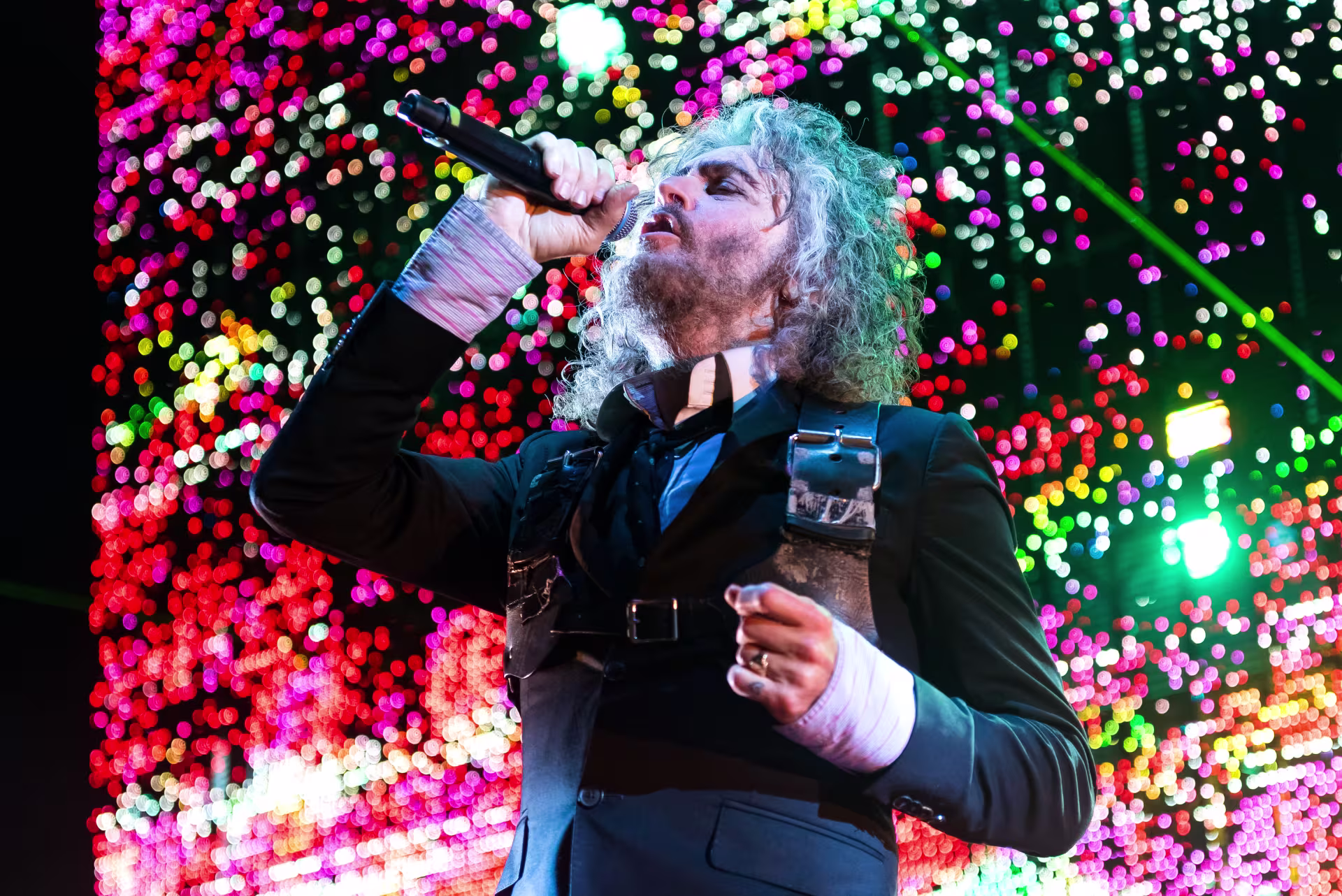A man with gray curly hair singing into a microphone with bright, red, pink, and yellow lights behind him.