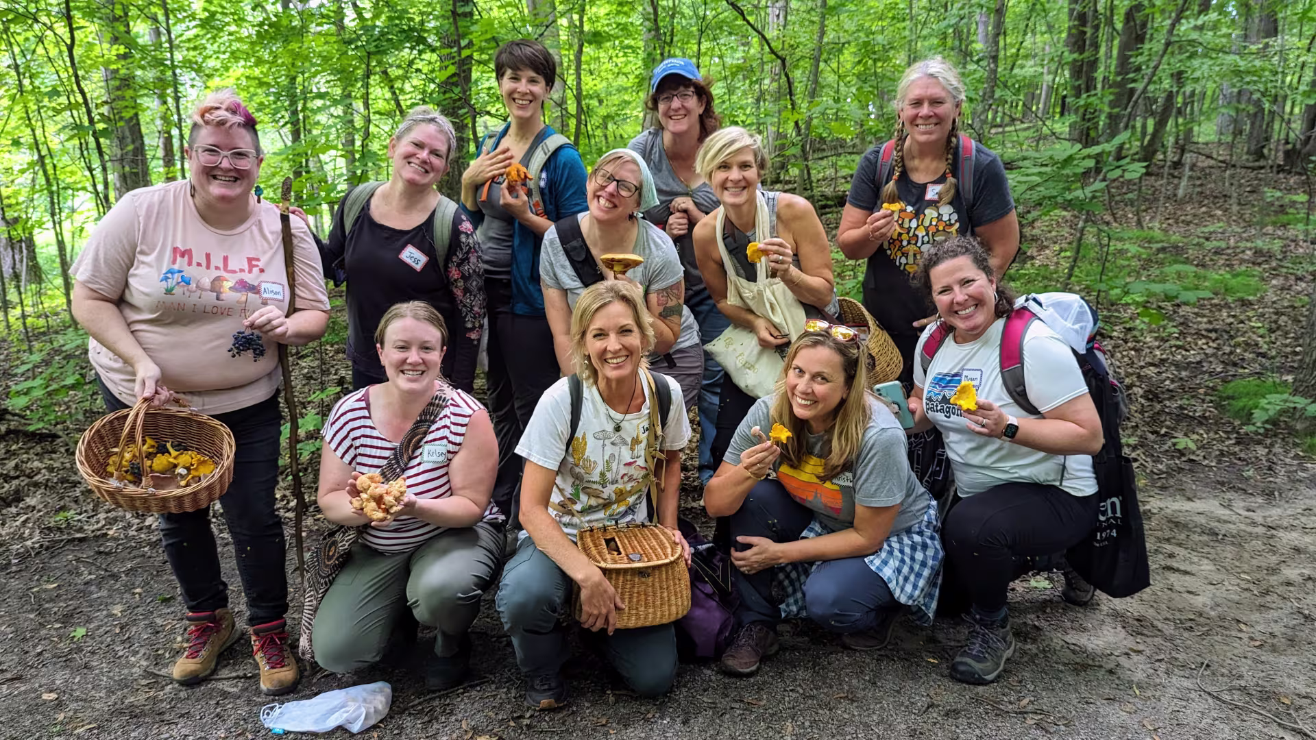 In an outdoor backdrop of forest trees, eleven foragers smile while holding individual mushrooms or baskets of mushrooms. Some wear backpacks.