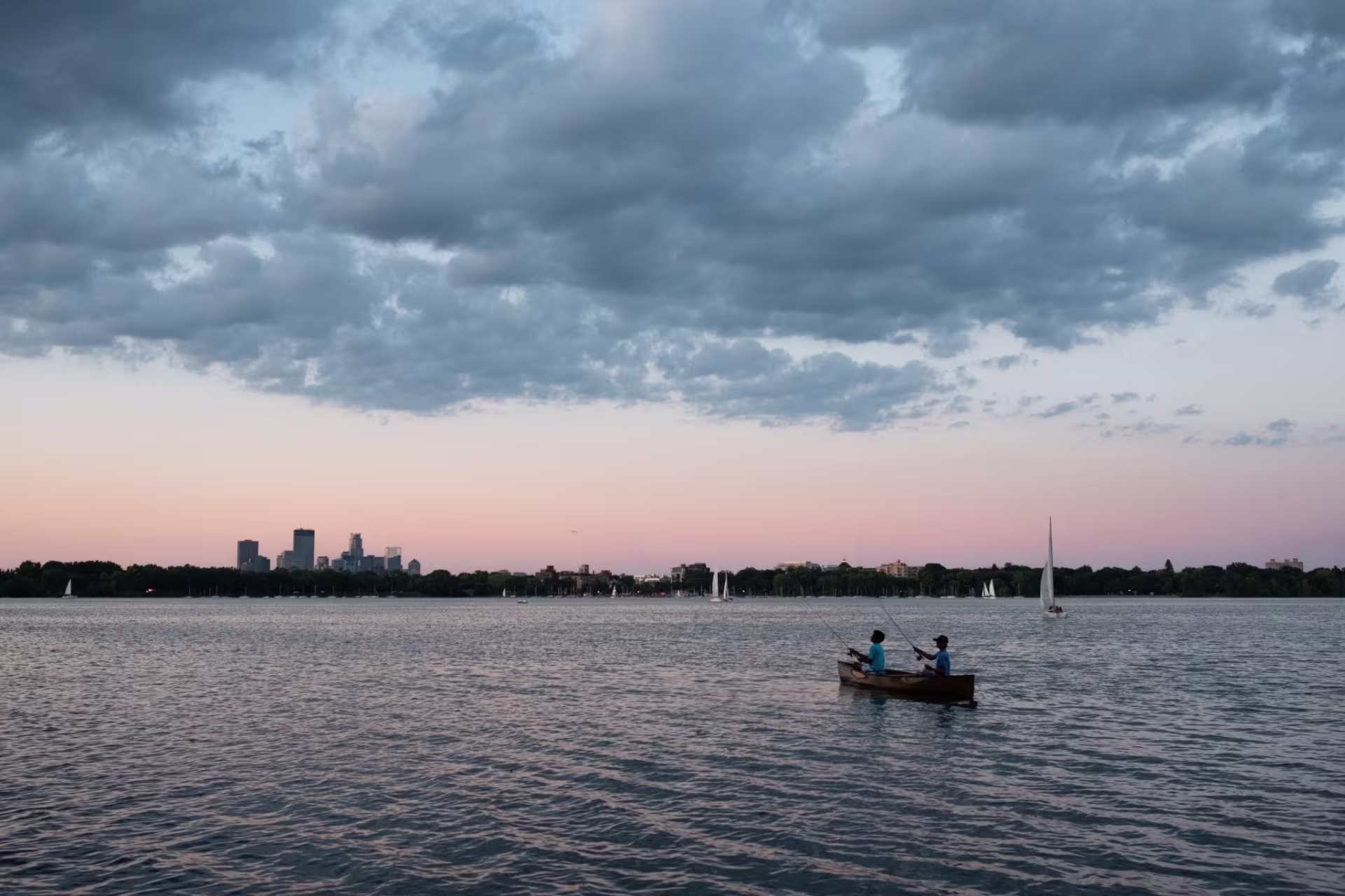 A cloudy sky during sunset. The minnesota skyline is in the background. The lake is on the bottom half of the image. There is a boat with two people fishing on it