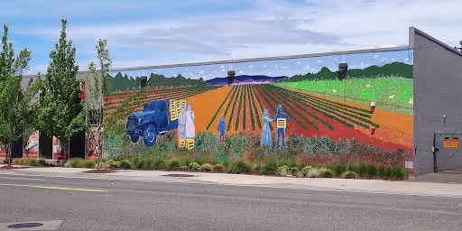 A mural at 10721 NE Sandy Blvd. in Portland’s Parkrose neighborhood depicts farmers working in fields colored orange, red, and green. The blue-hued figures load yellow crates onto a blue truck.