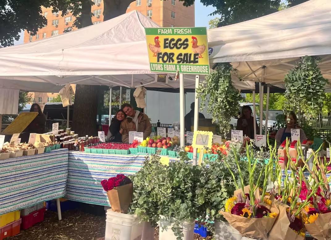 Two people posing for a photo at their booth. There is a sign that says "eggs for sale" and an assortment of flowers, and fruit at the stand
