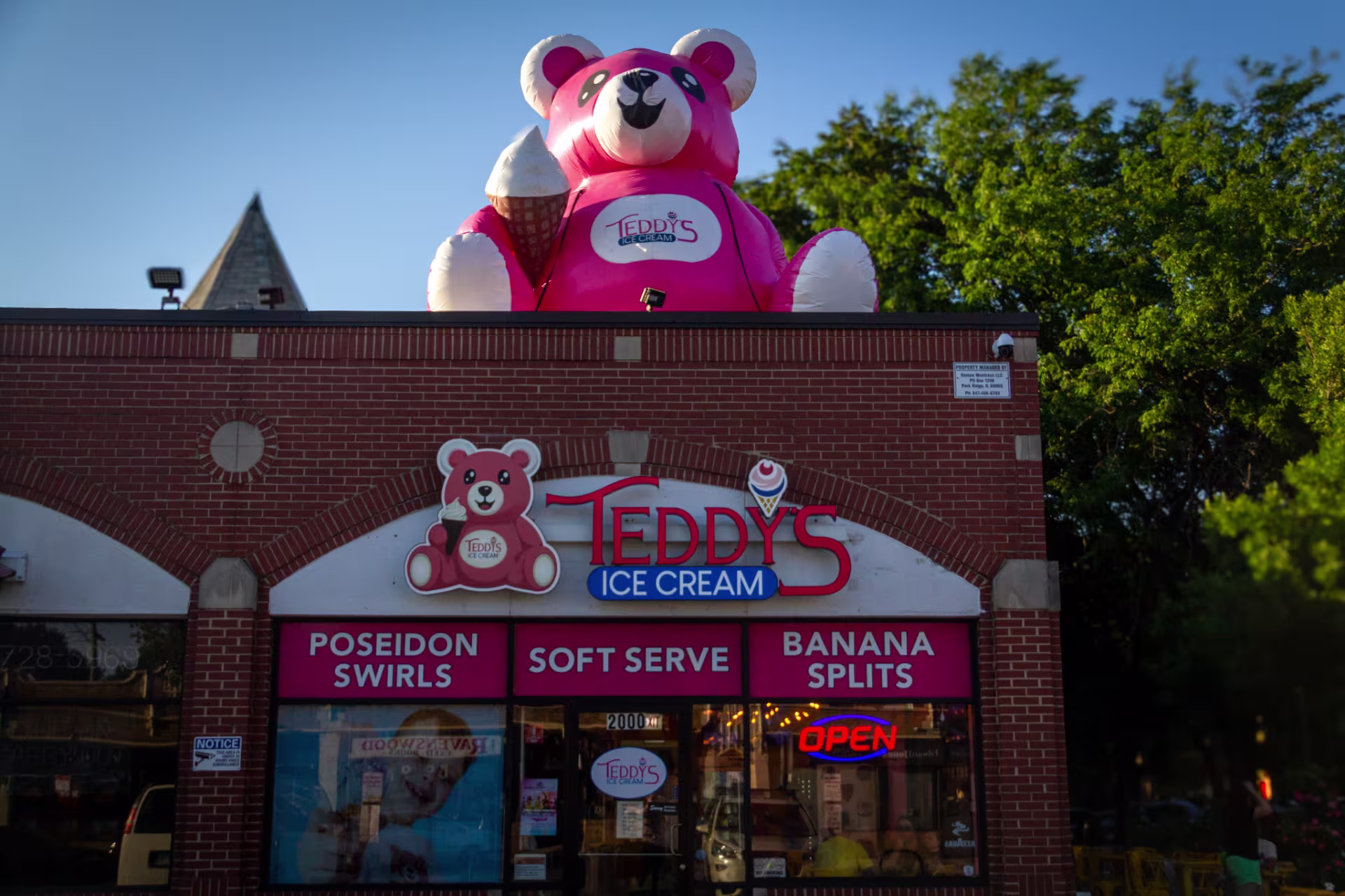 A brick one story building with a sign that says "teddy's ice cream" Next to the sign is a teddy bear eating ice cream. Under this sign there are three window panes with words that say "poseidon swirls" "soft serve" and "banana splits" On the roof there is an inflatable pink teddy bear with ice cream