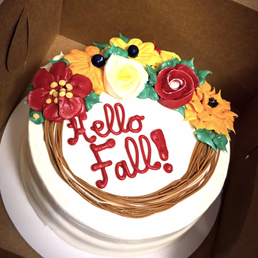 White cake inside a box with orange writing that says “Hello Fall!” and red and yellow flowers with green leaves made from icing.