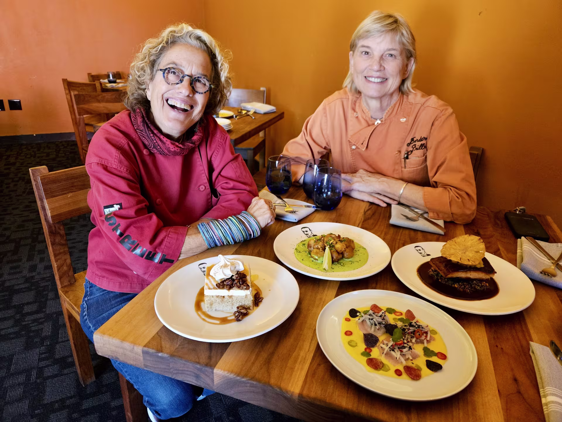 Susan Feniger and Mary Sue Milliken sit at a wooden table with four plates of different Mexican dishes.