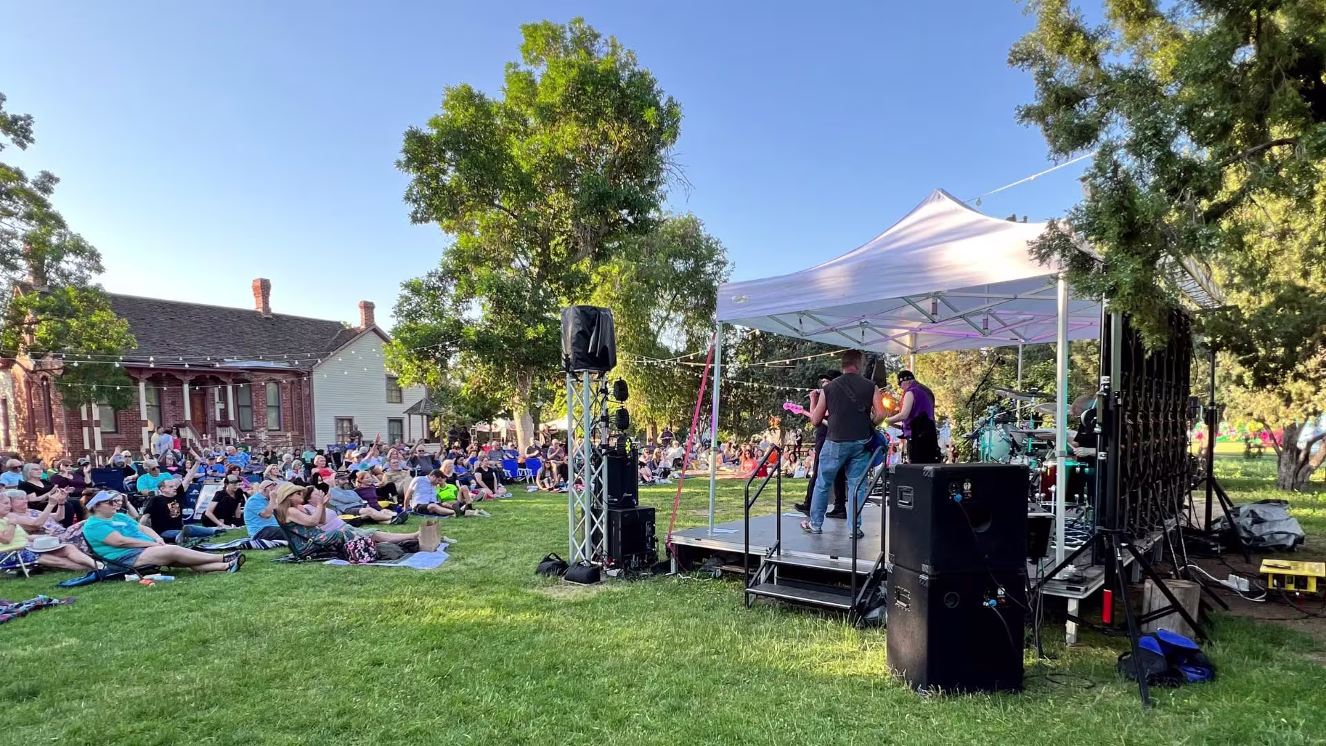 Outdoor performers under a white tent perform on stage while concertgoers sit on green grass listening. A brick building is behind the concertgoers to the left, and string lights run parallel to the crowd and stage.