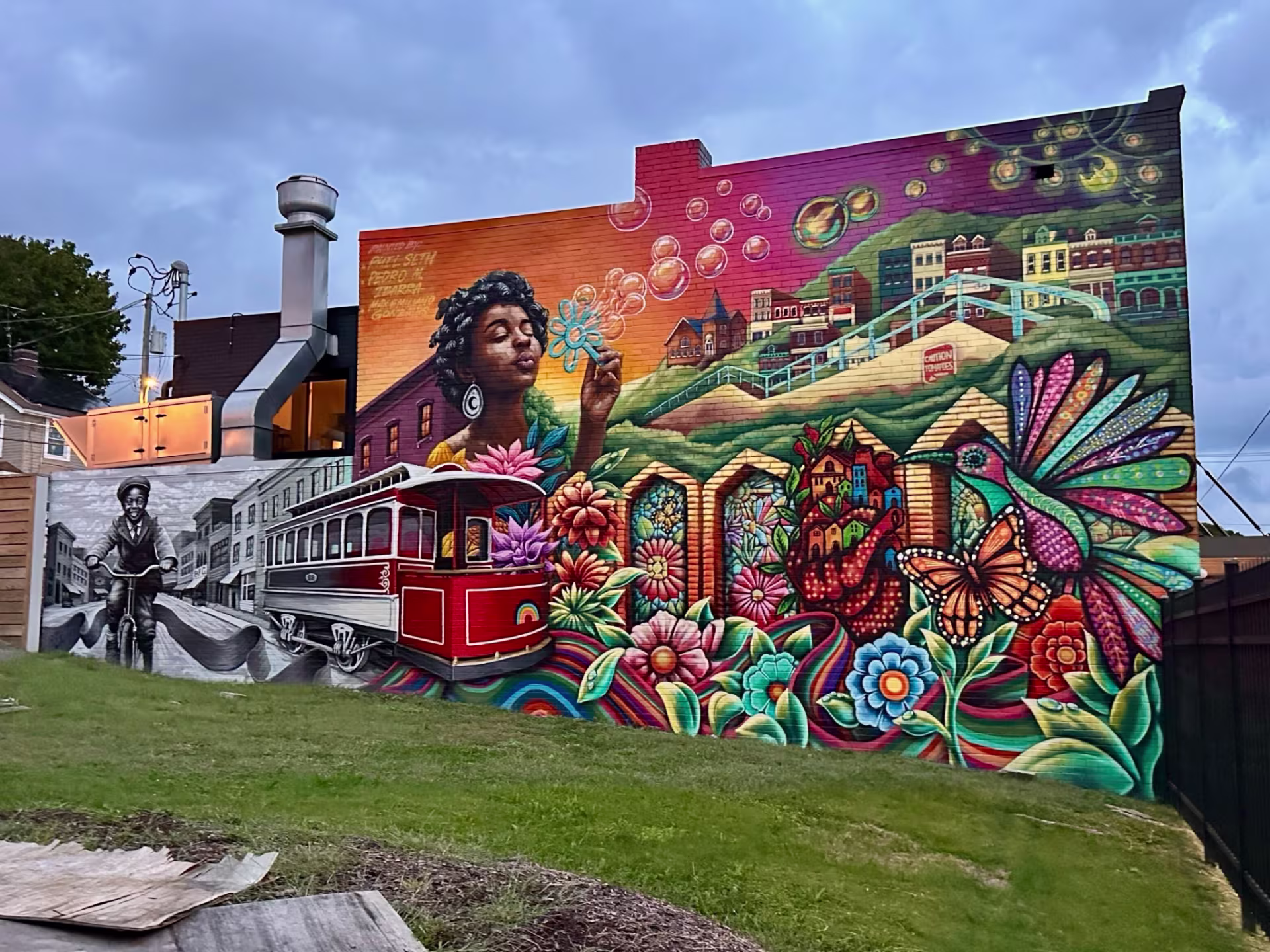 A colorful mural on the side of a building includes depictions of a street car, a woman blowing bubbles, and a humming bird.