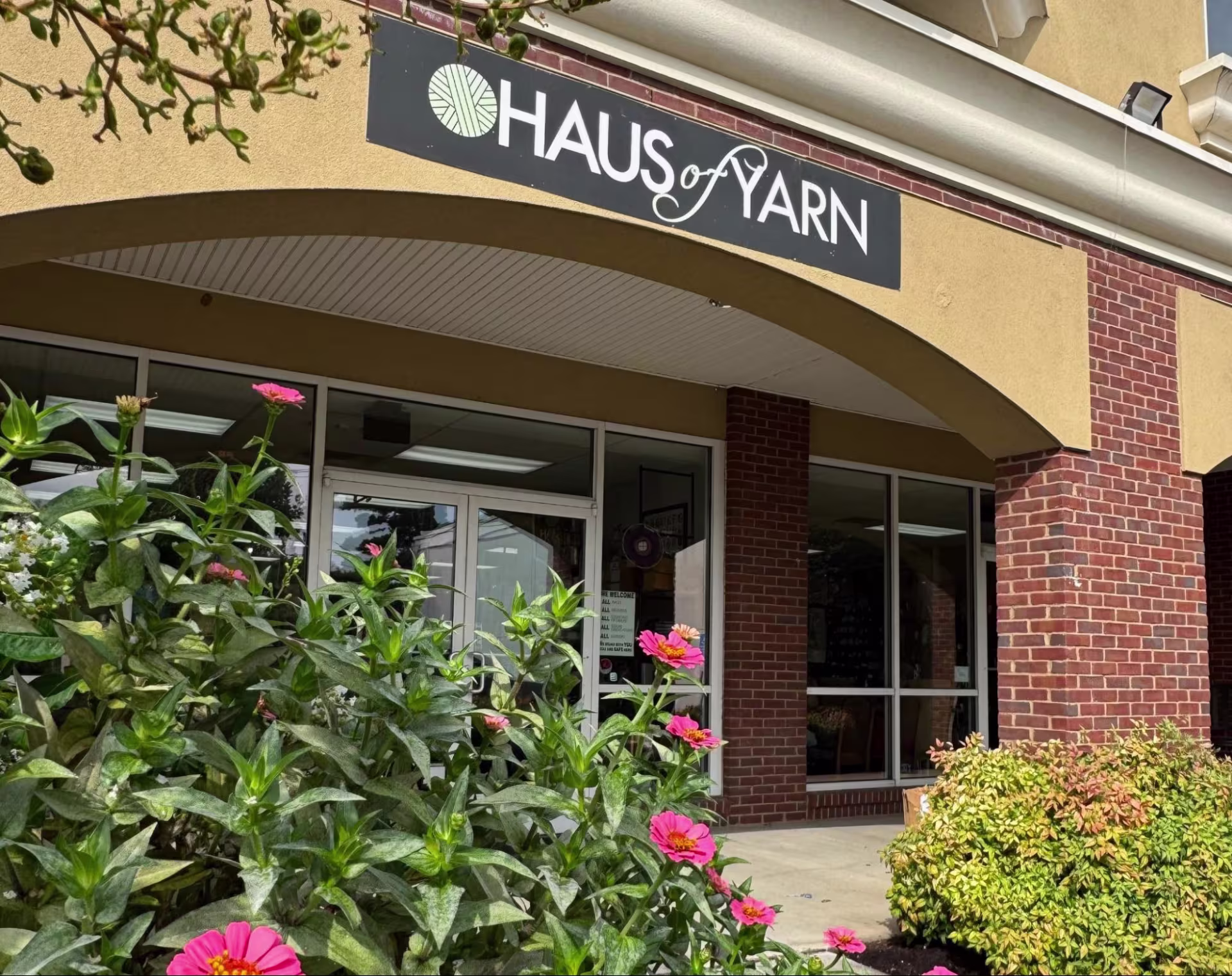 A modern storefront on White Bridge Road features an arched entrance supported by brick columns. A sign reading “Haus of Yarn” hangs above the arch and hot pink zinnias bloom in front.