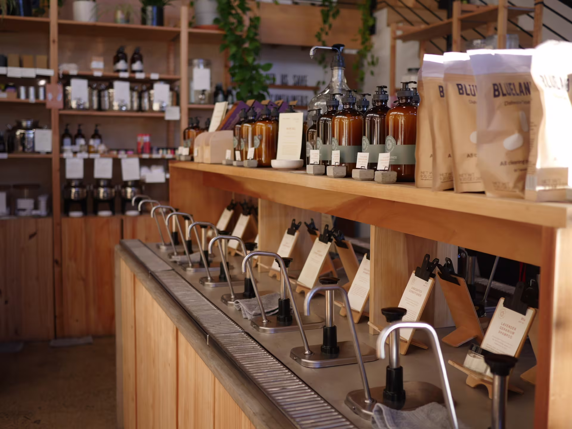 A room lined with wooden shelves. In the foreground is a counter with metal pump spouts over a metal grate that dispense various products. Above them are glass bottles of samples to try.