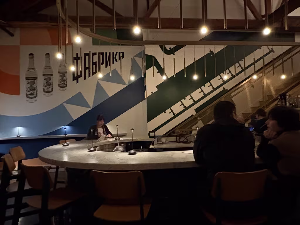 horse shoe of a bar with patrons around it with cyrillic writing and and bottles feature on the wallpaper