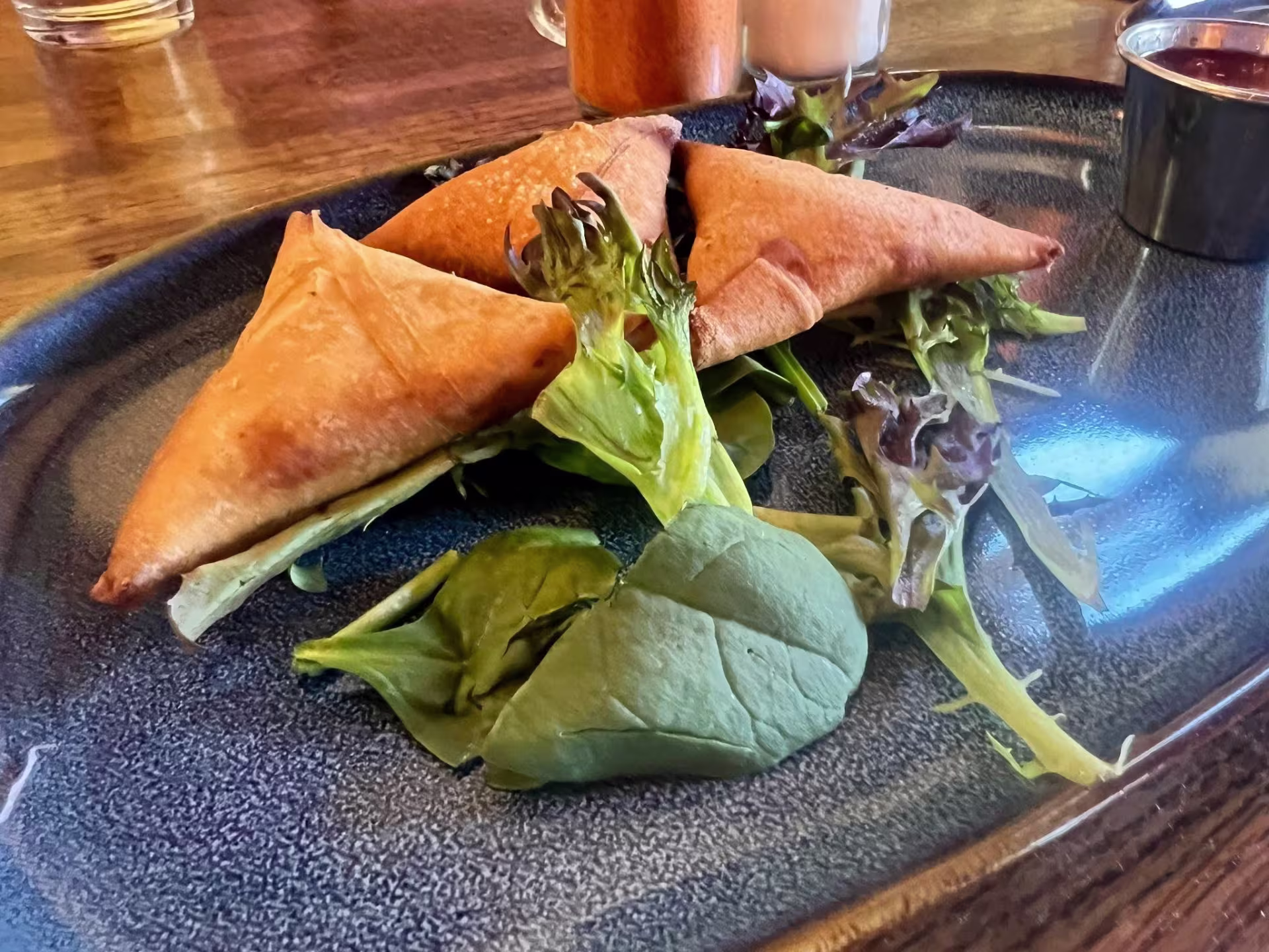 Three sambusas sitting on an oval blue plate with some greens and a dipping sauce in a metal container