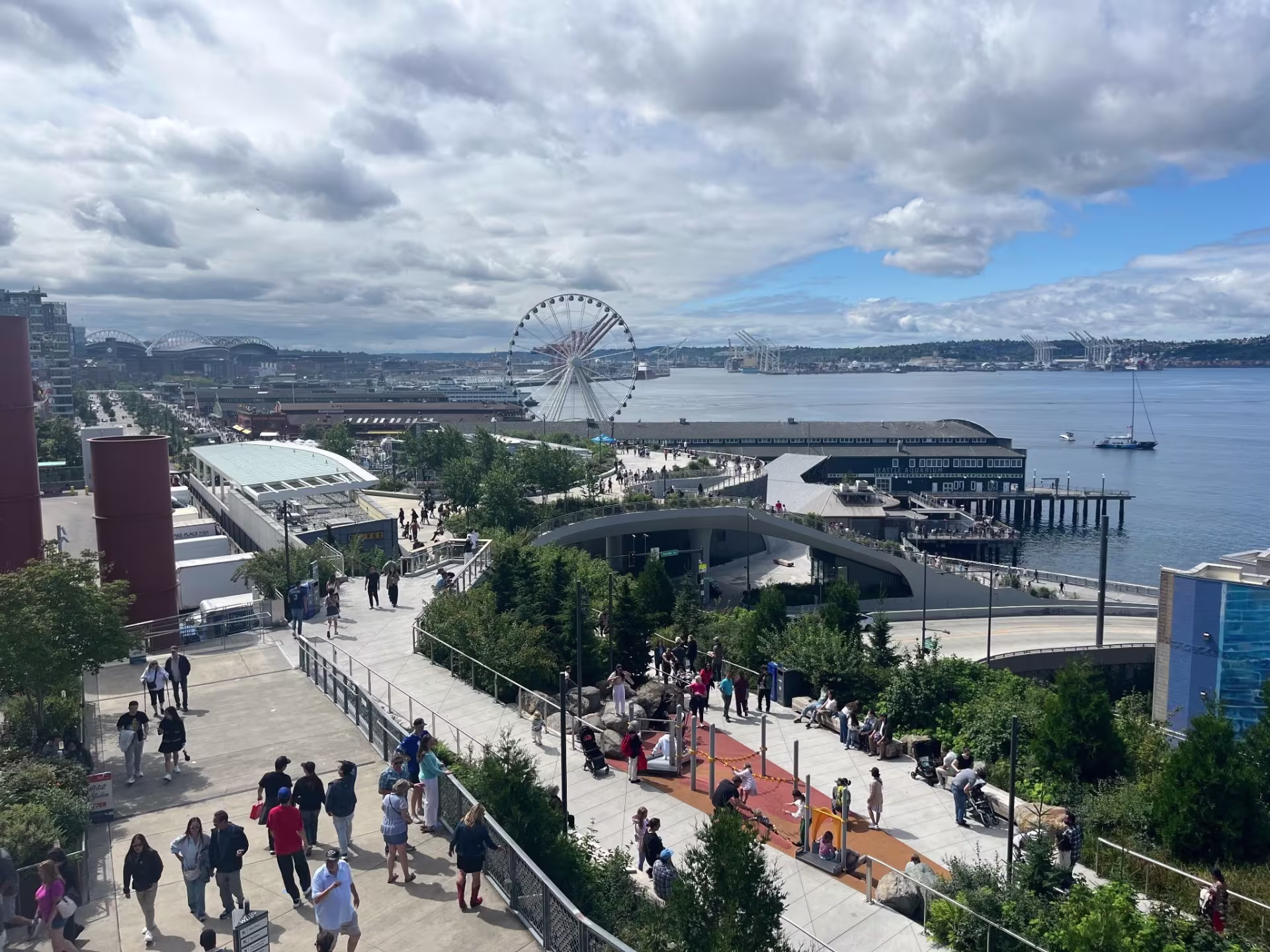 A view of the Seattle waterfront from a higher floor. The sky is lightly cloudy, people are walking and in the back there is a view of the ferris wheel