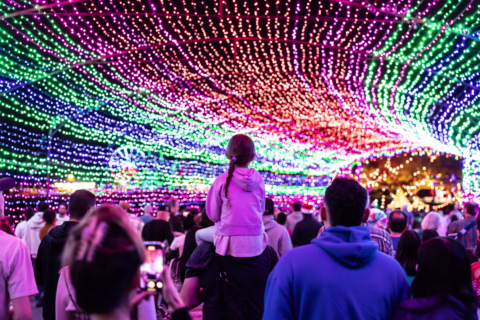 A child on someone's shoulders looking at a tunnel of Christmas lights. 