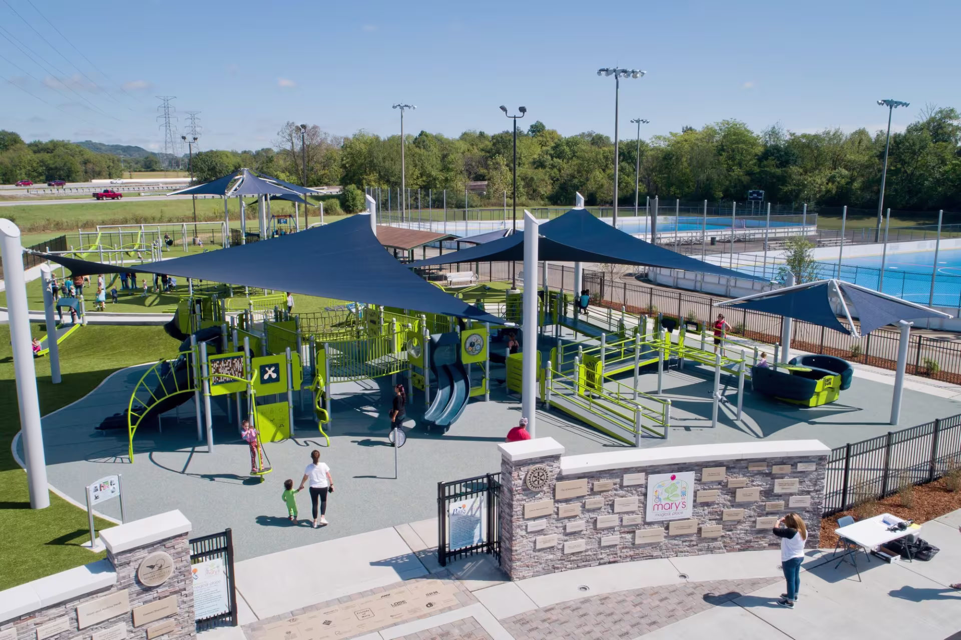 Aerial view of an inclusive playground featuring accessible play structures in lime green and navy under large fabric shade sails. Ramps with railings lead up to elevated platforms with slides, and a sensory swing and accessible roundabout sit nearby on rubberized safety surfacing. Families with children walk through the space, with a memorial donor wall and entrance gate in the foreground, an outdoor pool and roller hockey rink in the background, and a tree line and highway visible in the distance under a clear blue sky.