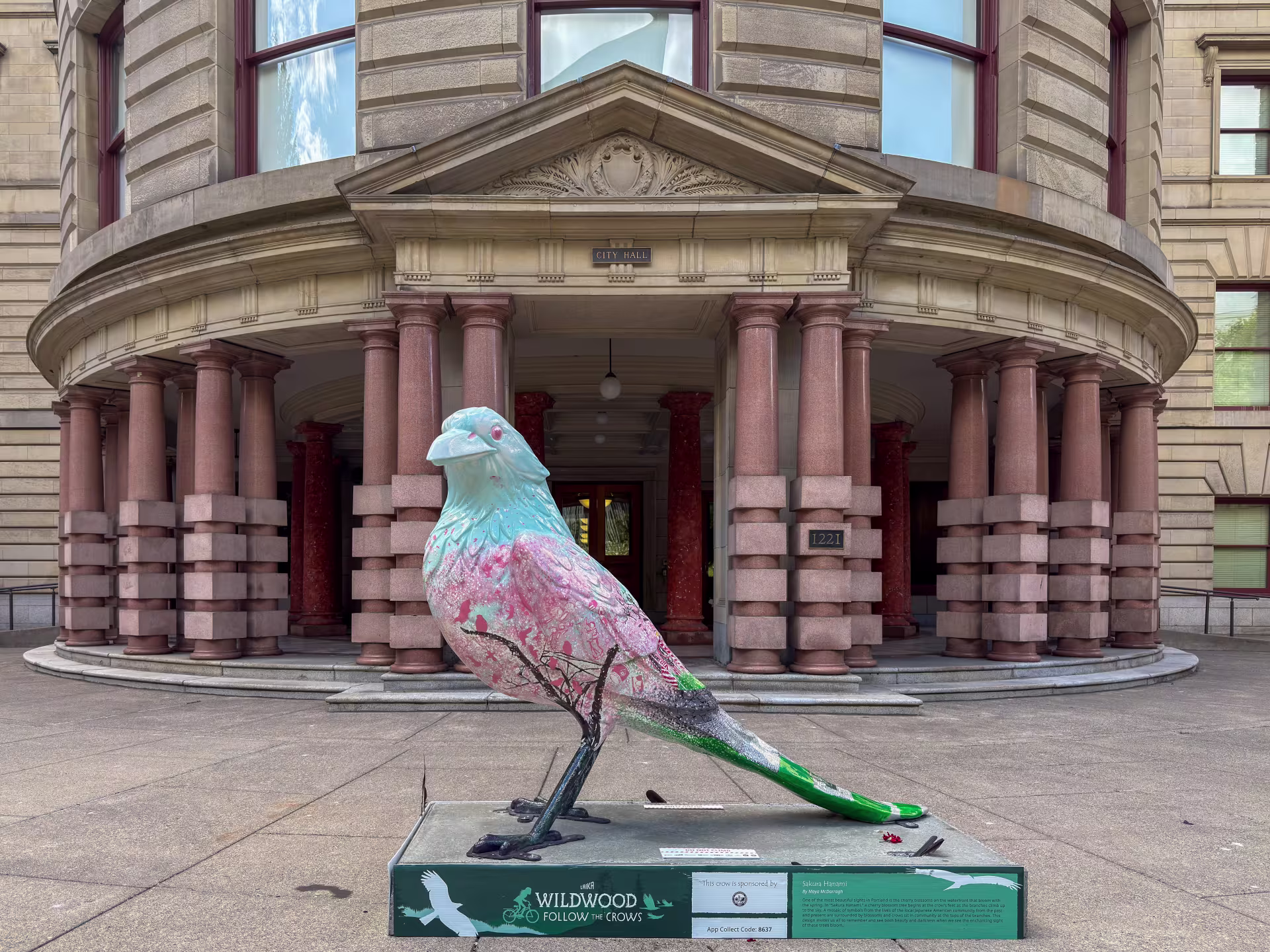 A crow statue painted with a light blue head and a body with blossoming cherry trees sits out front of Portland City Hall. The installation is part of the Wildwood: Follow the Crows temporary art trail. 