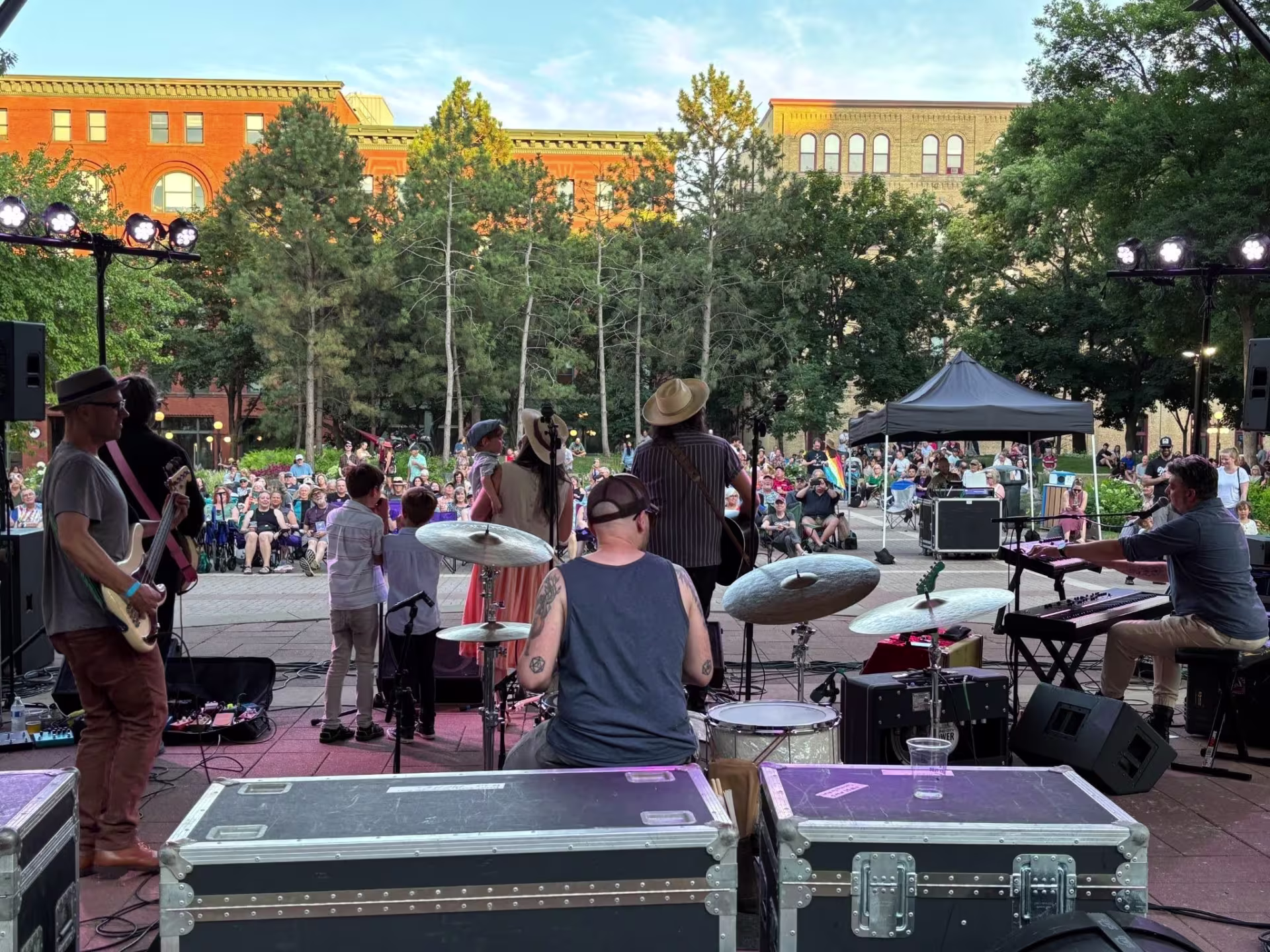 Band plays to audience in park, as viewed from behind the band. Brick buildings and trees in background.