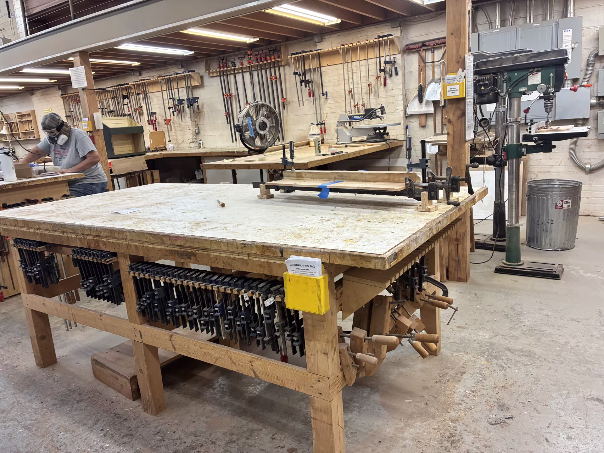  Inside the wood shop at Philadelphia Woodworks, with a woman working at one of the workshop tables in the background. A large table in the foreground is lined with dozens of clamps along its edges; walls are hung with organized rows of tools and clamps; there is a drill press in the corner.