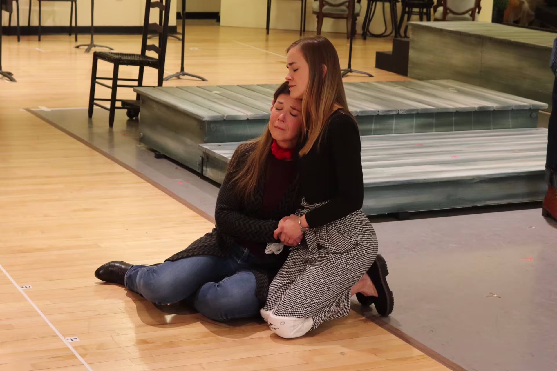 Two women sitting on a wooden floor hugging.