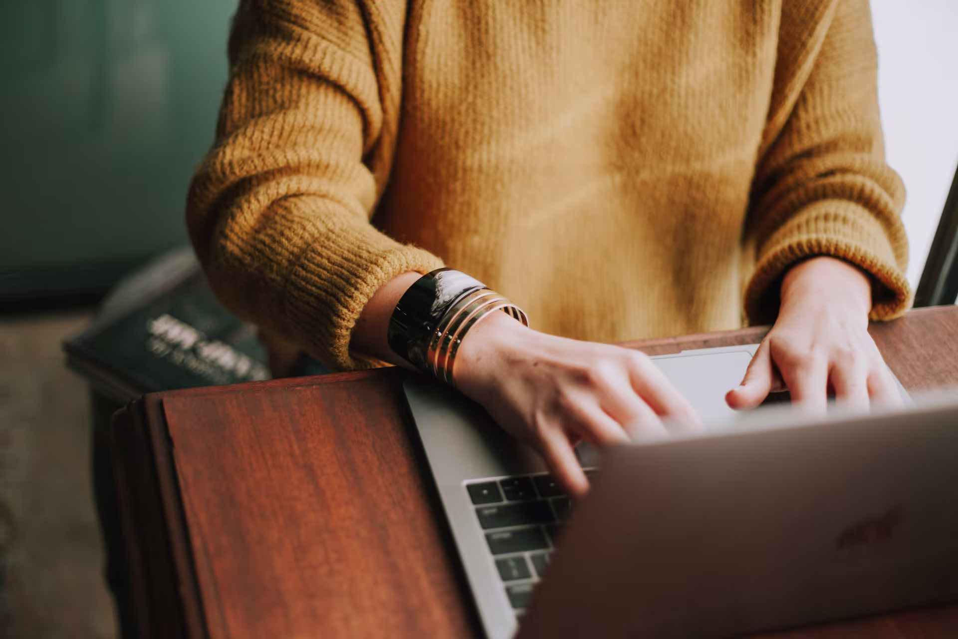 A woman Iight skinned with bracelets wearing a mustard sweater) types on a laptop that sits on a wooden desk.