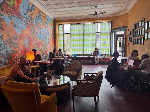 Interior of Eremita Cafe in Roxborough, with customers sitting at tables and vintage upholstered chairs beneath a pressed-tin ceiling. A colorful abstract mural covers the left wall, while framed artwork lines the opposite wall and tall windows fill the space with natural light.