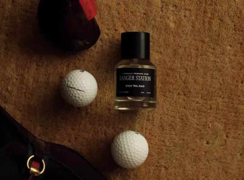 A bottle of Ranger Station cologne with a black label and lid lies on a brown surface next to a couple of white golf balls and a golf club.