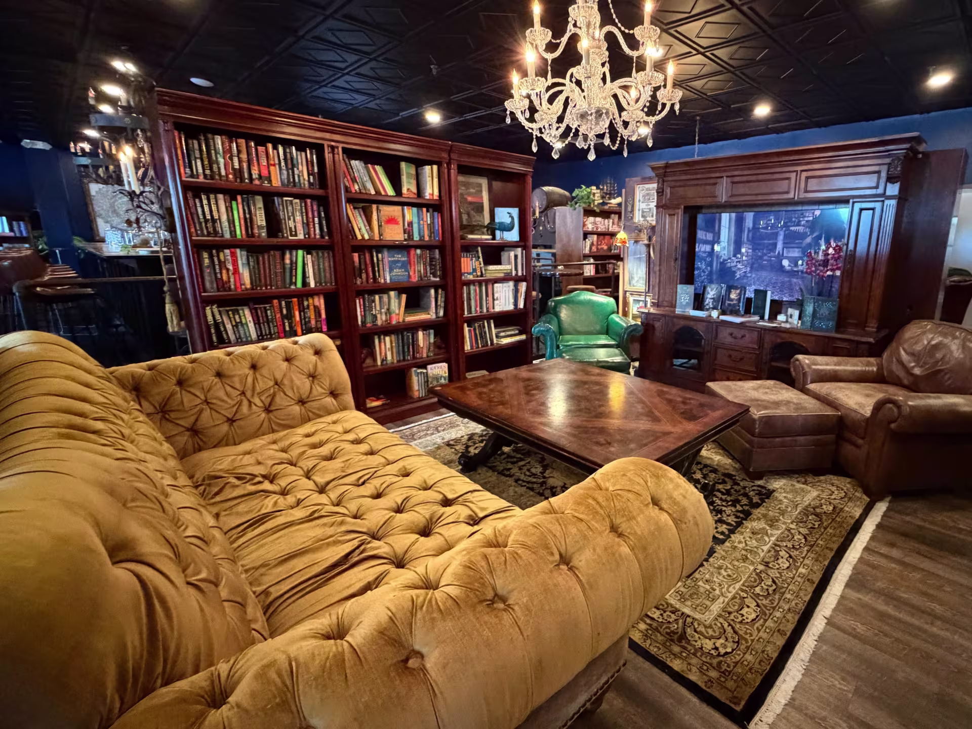 A moody reading room, with dark wood bookshelves and a coffee table surrounded by a giant golden tufted sofa and two leather armchairs. A chandelier hangs from the black tin ceiling.