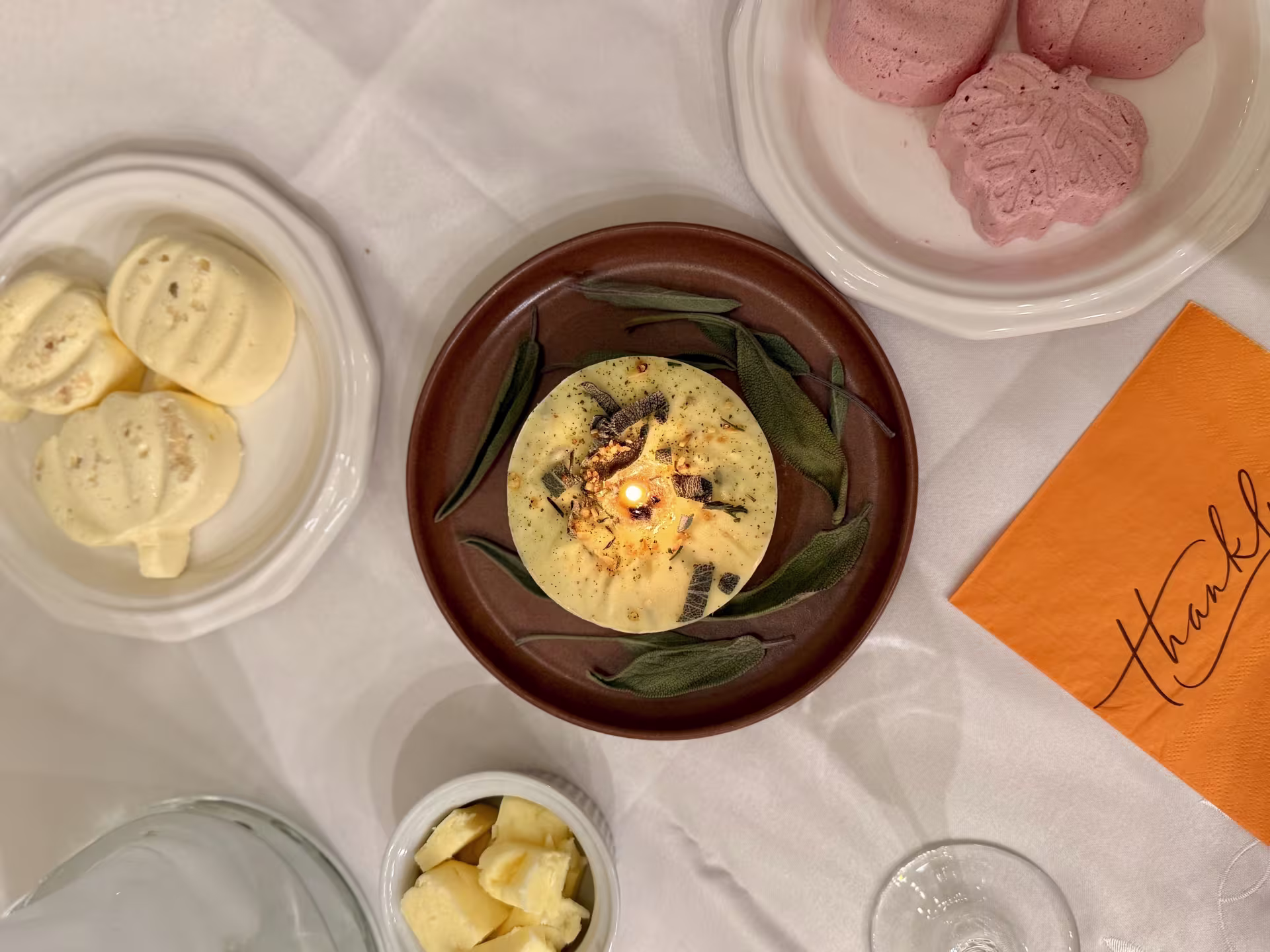  A yellow butter candle is the centerpiece of a tablescape, sitting on a red ceramic plate and surrounded by fall-themed cookies and an orange napkin that says, "thankful."