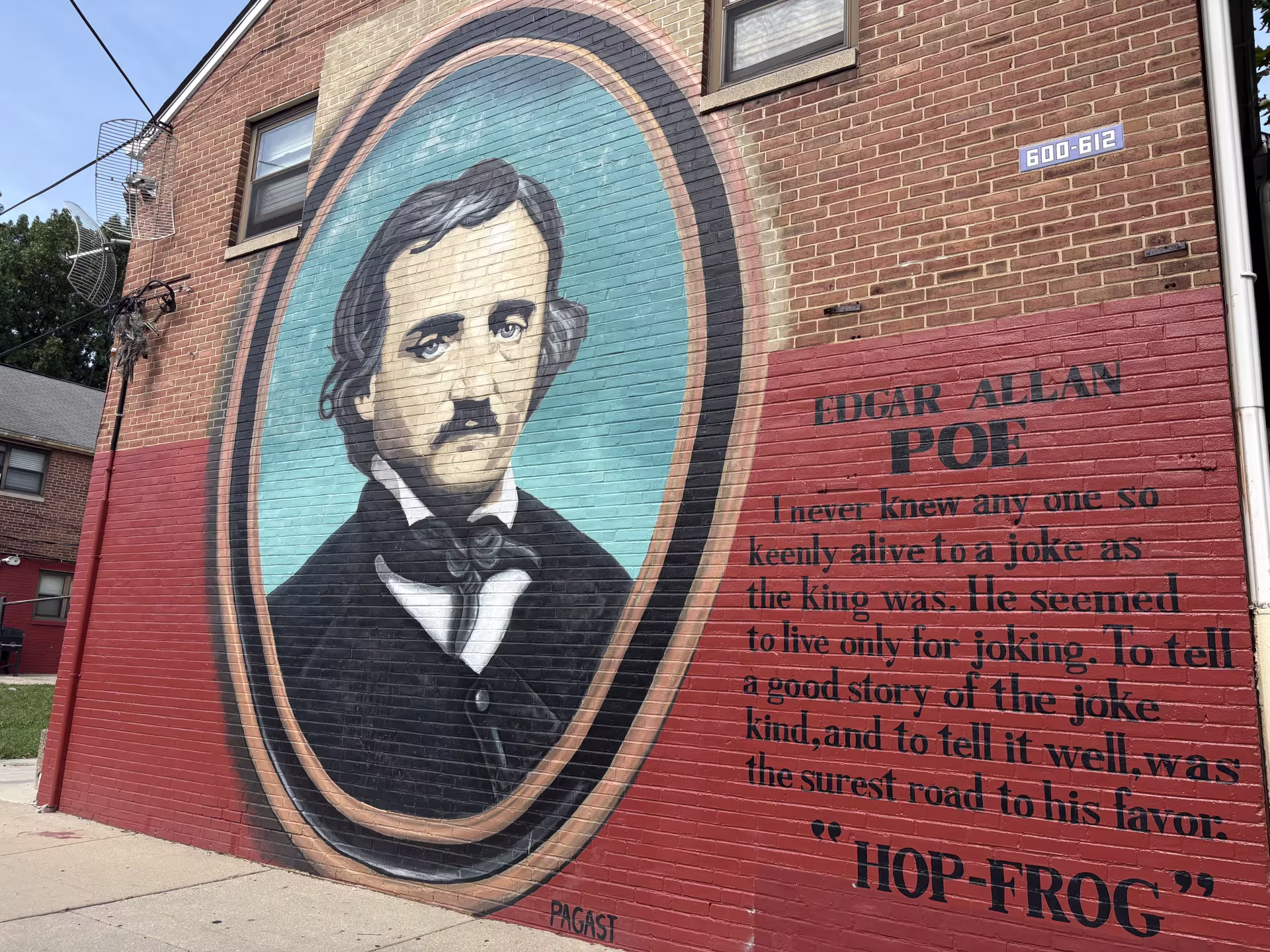 A mural on a brick wall depicting a portrait of Edgar Allan Poe against a blue background in an oval frame. He is wearing a black dress coat and tie, and white shirt. Beside him is a quote from his short story Hop-Frog: “ I never knew anyone so keenly alive to joke as the king was. He seemed to live only for joking. To tell a good story of the joke kind, and to tell it well, was the surest road to his favor.” 