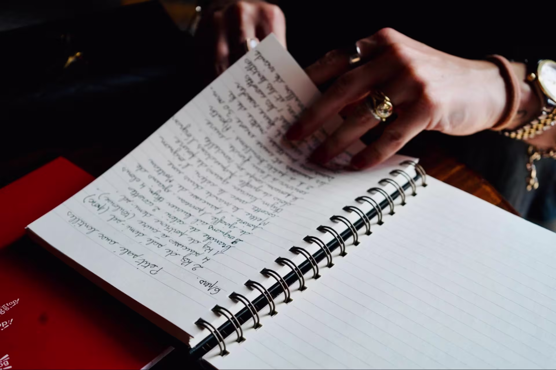 A hand wearing rings flips through a spiral-bound, lined notebook containing handwritten recipes in French. 