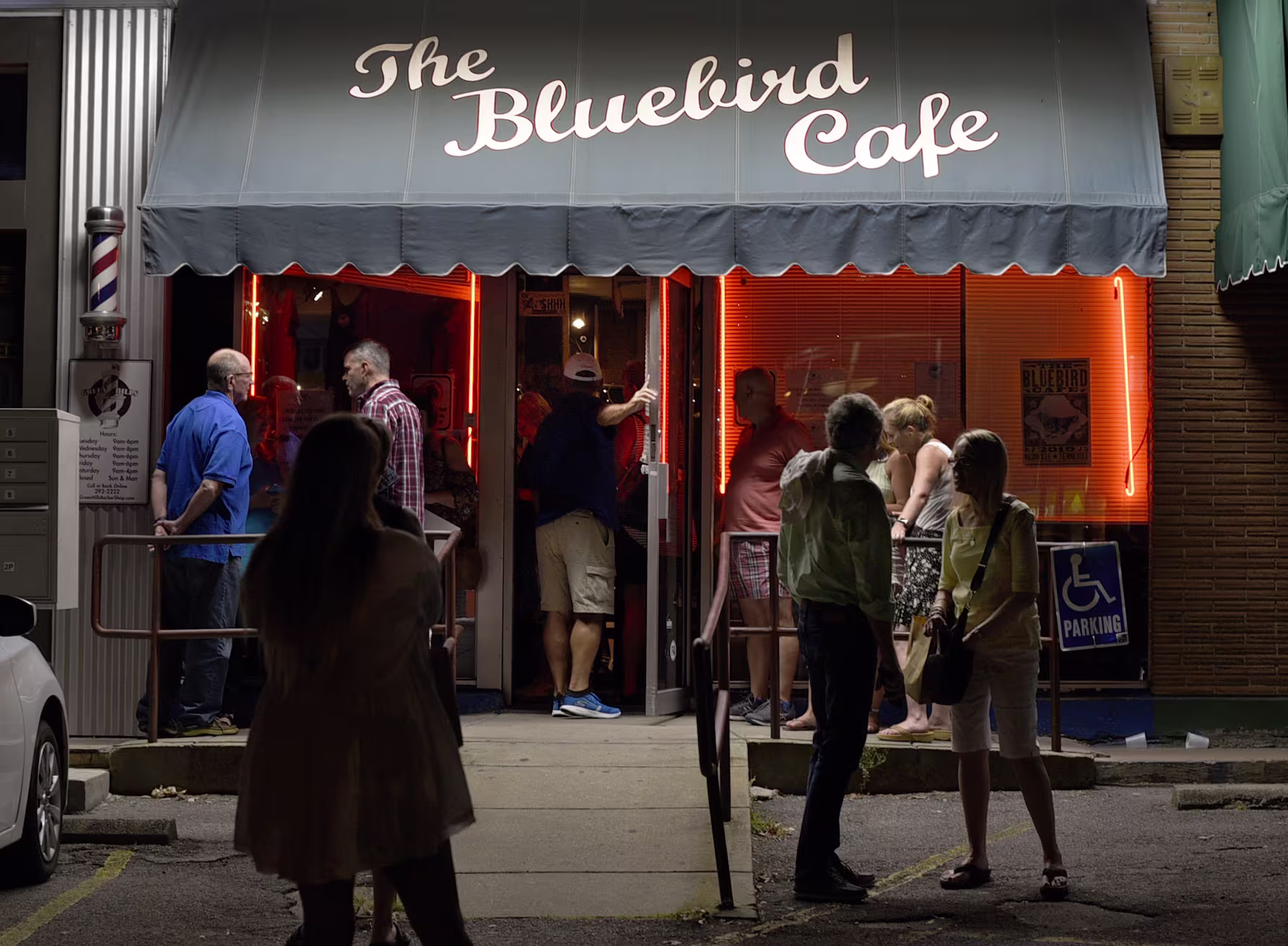 People gather outside the Bluebird Cafe at night, waiting to enter the iconic Nashville music venue. A teal awning with the café’s name hangs above the entrance, red neon lights glow through the windows, and small groups chat on the sidewalk near the door.