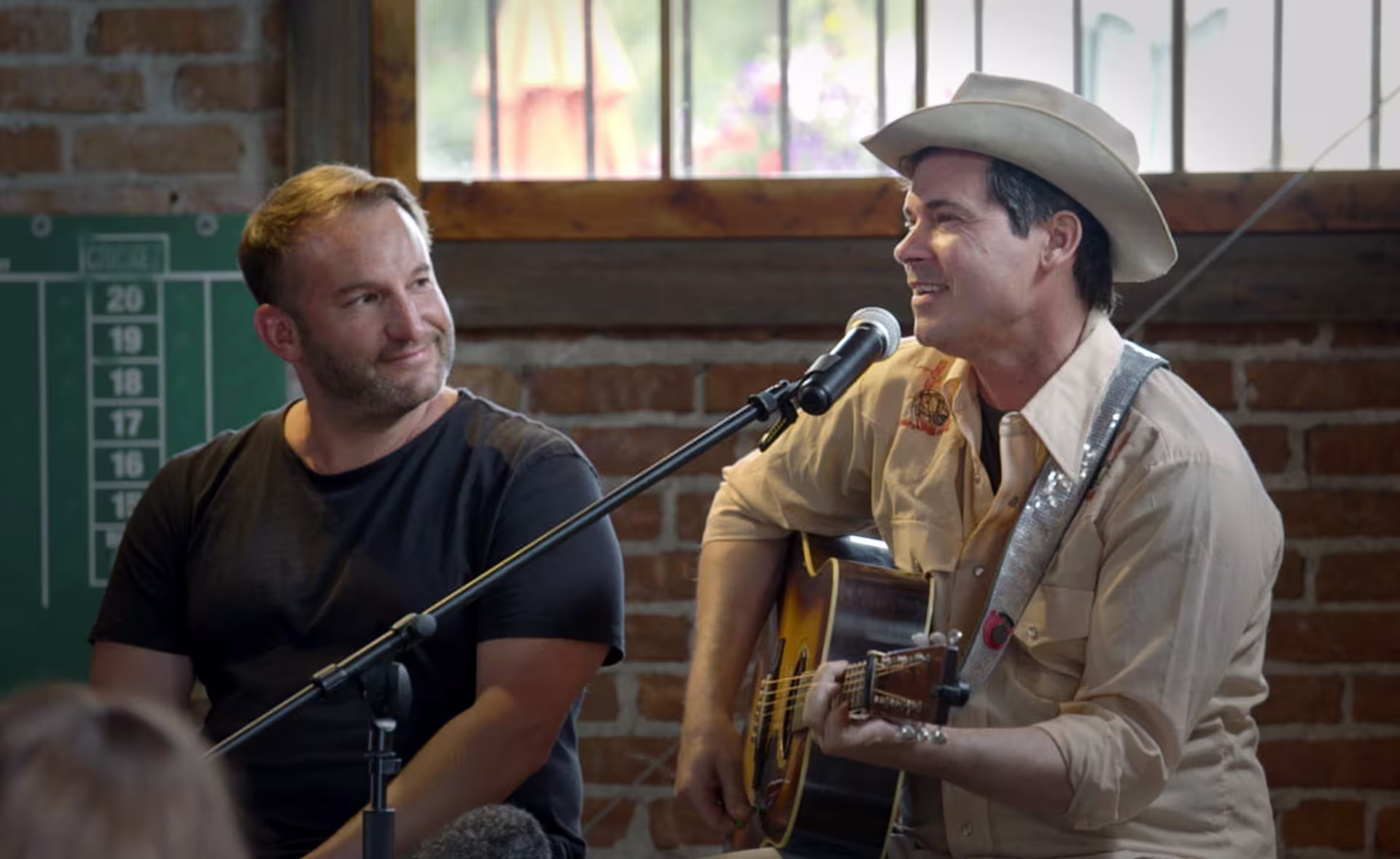 Two white men on a stage. One wears a black tee and is smiling at the second, who has a guitar and is speaking into a mic. He's wearing a tan button-up and a tan cowboy hat.