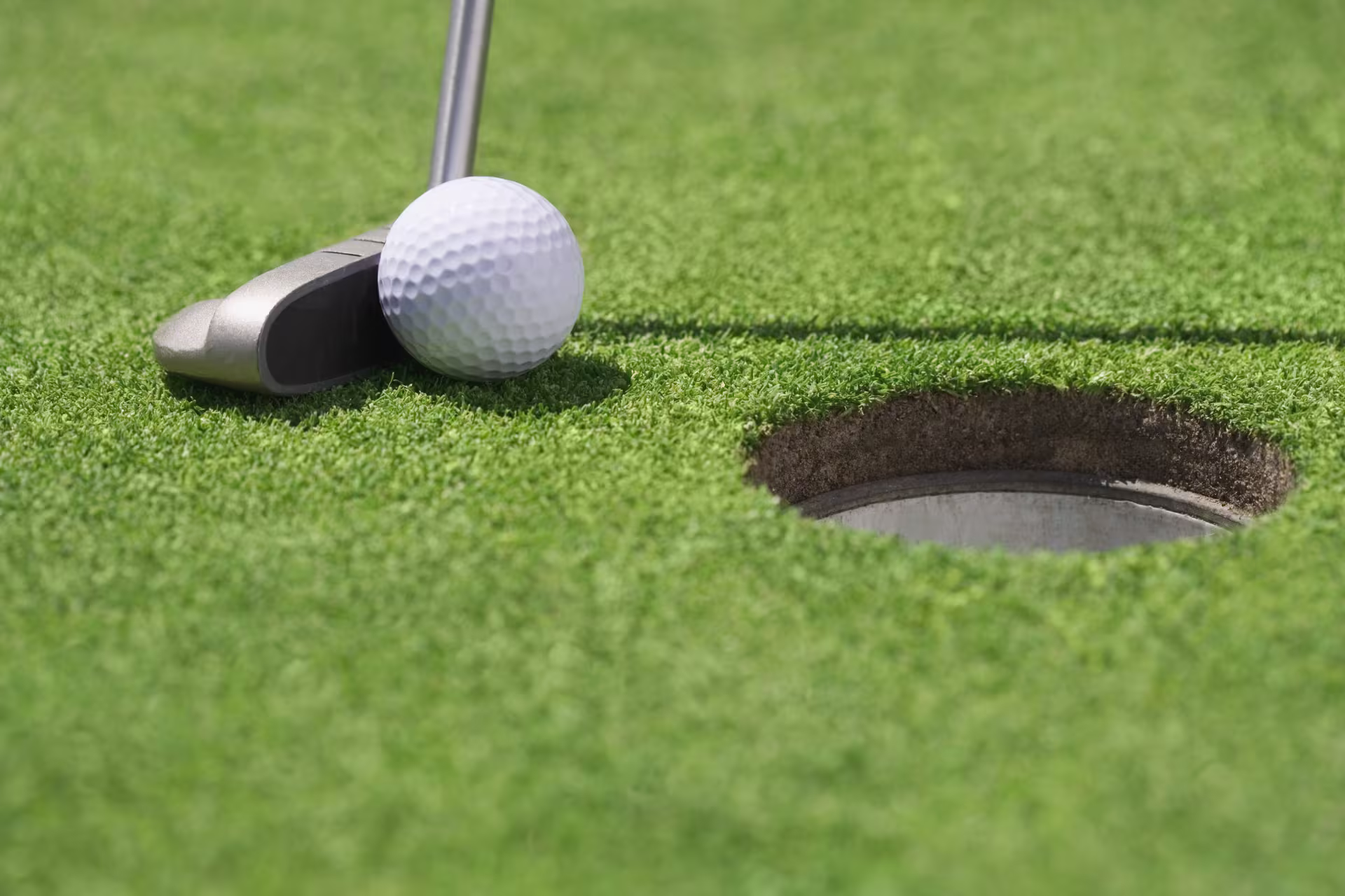 A green golf course with a white golf ball and a club next to a hole.