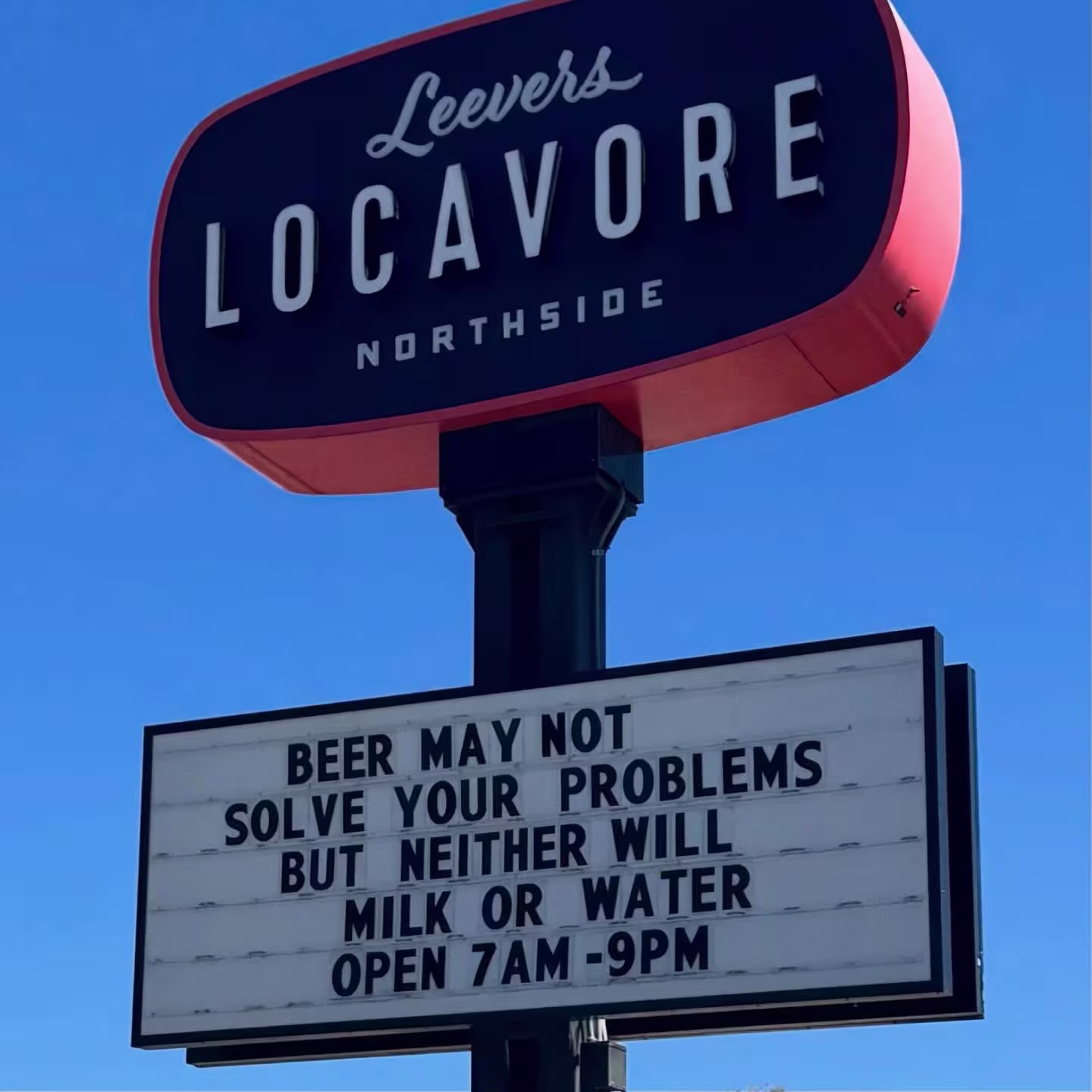  Sign outside of grocery store with a funny saying that reads, “Beer may not solve your problems, but neither will milk or water. Open 7 AM - 9 PM,” and blue sky in the background.