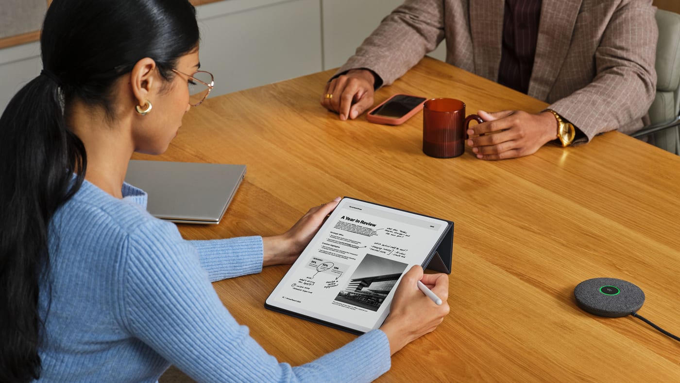 Mulher faz anotações à mão com a caneta no Kindle Scribe, sobre um documento aberto na tela, numa mesa de reunião