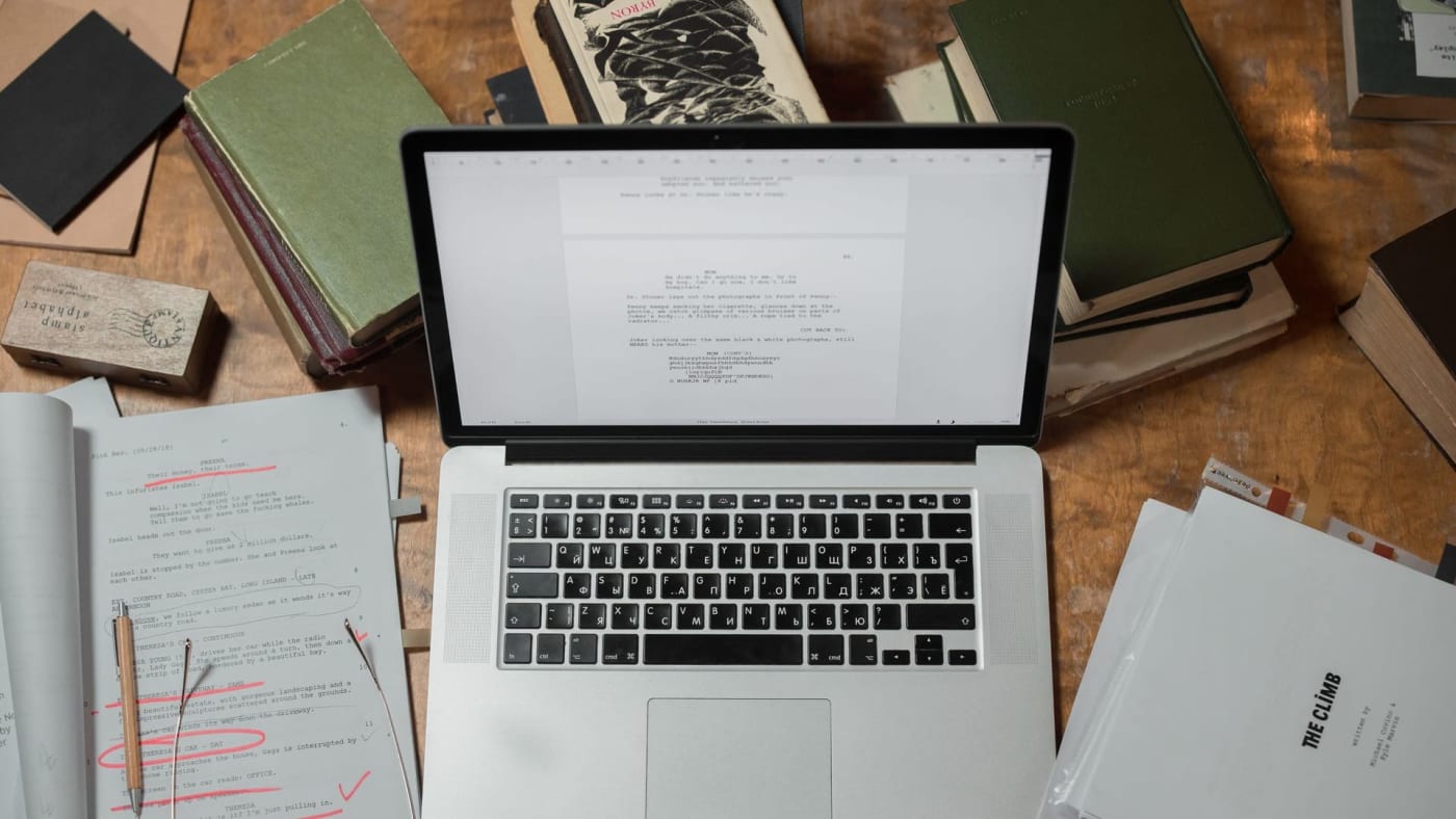 Mesa de trabalho vista de cima com laptop aberto exibindo texto, folhas com anotações e vários livros ao redor.