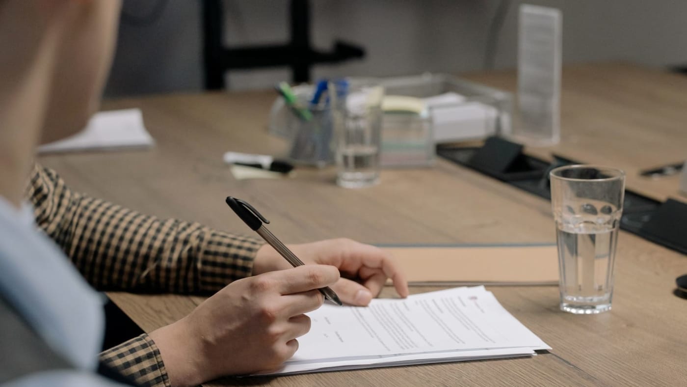 Pessoa segurando uma caneta enquanto analisa um contrato sobre uma mesa de escritório.