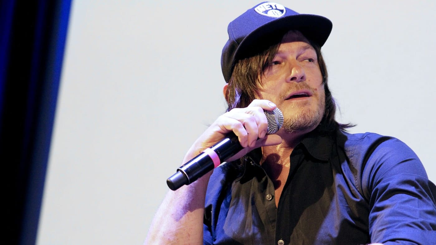 Norman Reedus falando ao microfone durante um evento usando boné preto