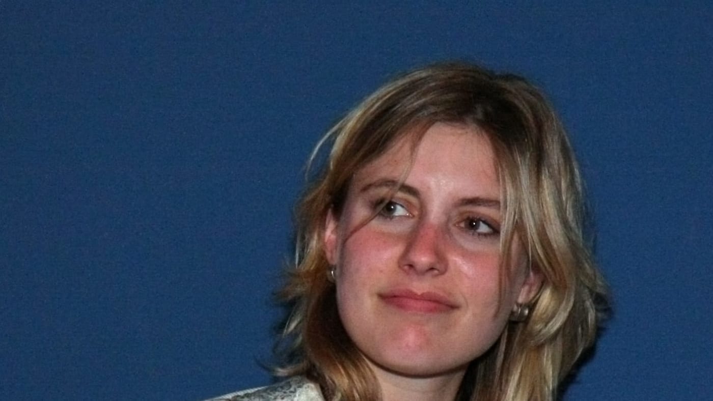 Greta Gerwig com vestido floral e cardigã branco contra fundo azul