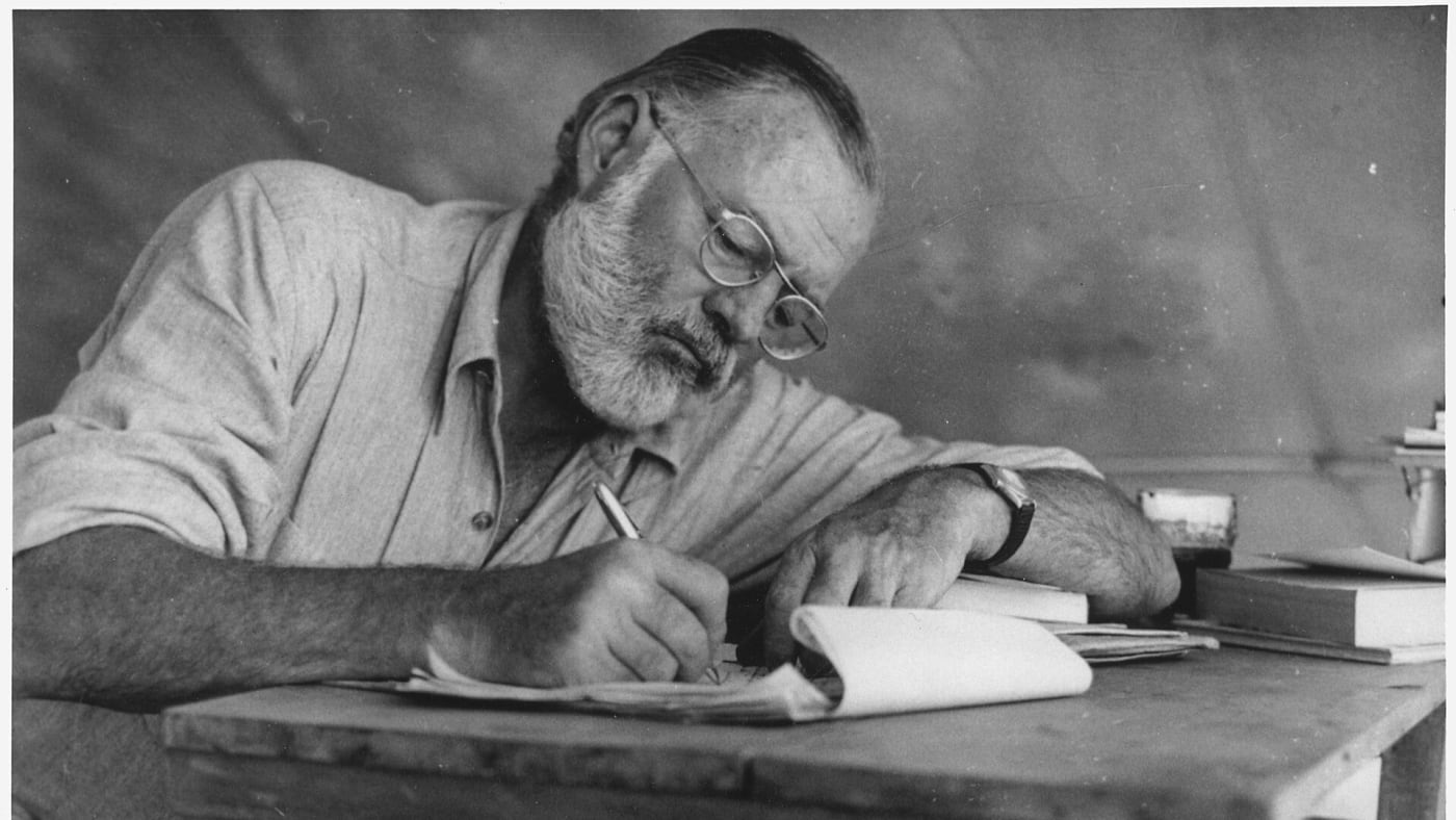 Ernest Hemingway escrevendo apoiado em uma mesa rústica